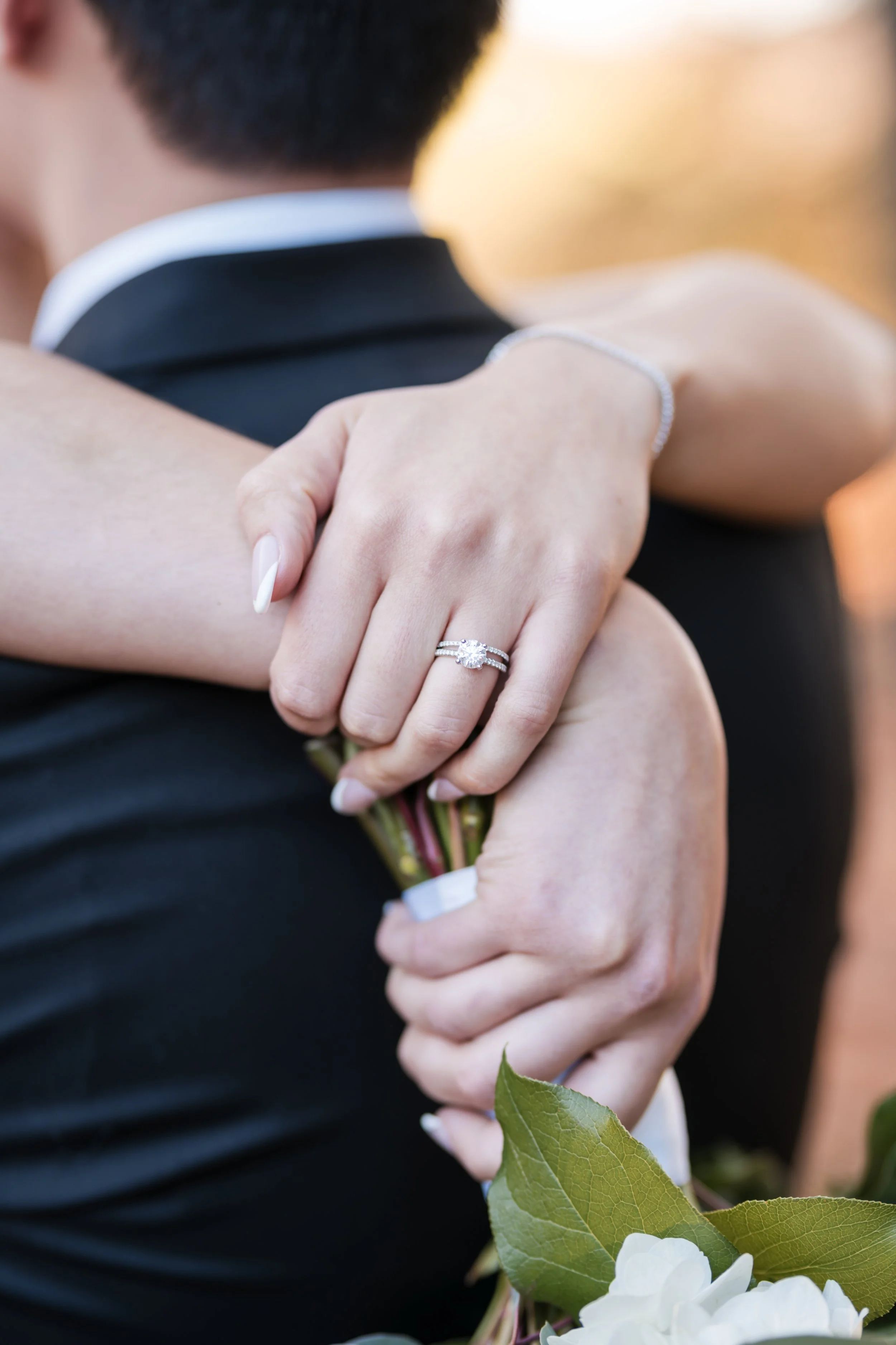Close-up of a couple hugging, with focus on the woman's hand showing an engagement ring and wedding band, holding a bouquet of white flowers with green leaves.