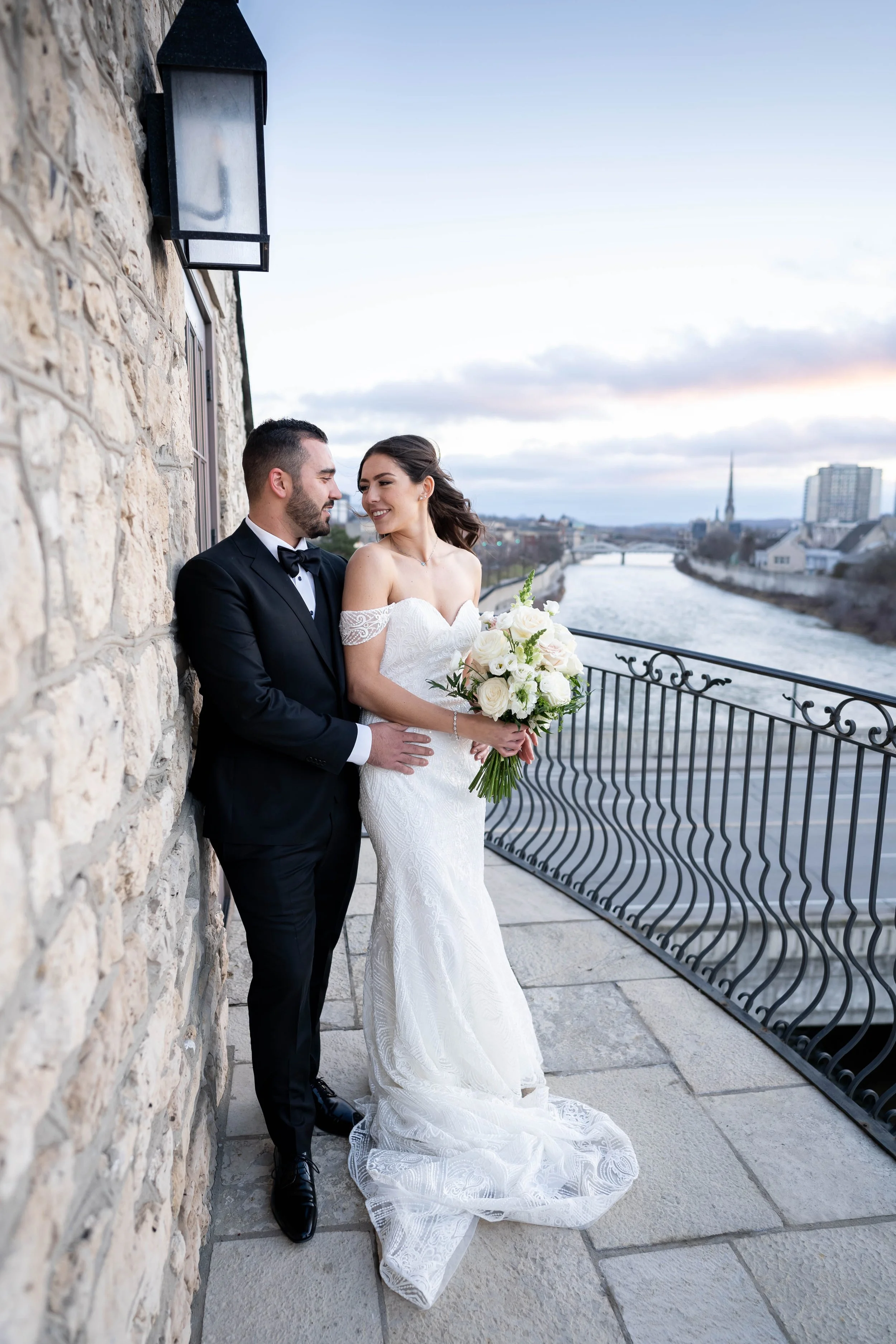 Bride and groom standing on a balcony overlooking a river, with city buildings in the background, during sunset. The bride is holding a bouquet of white roses, and the couple is smiling at each other.