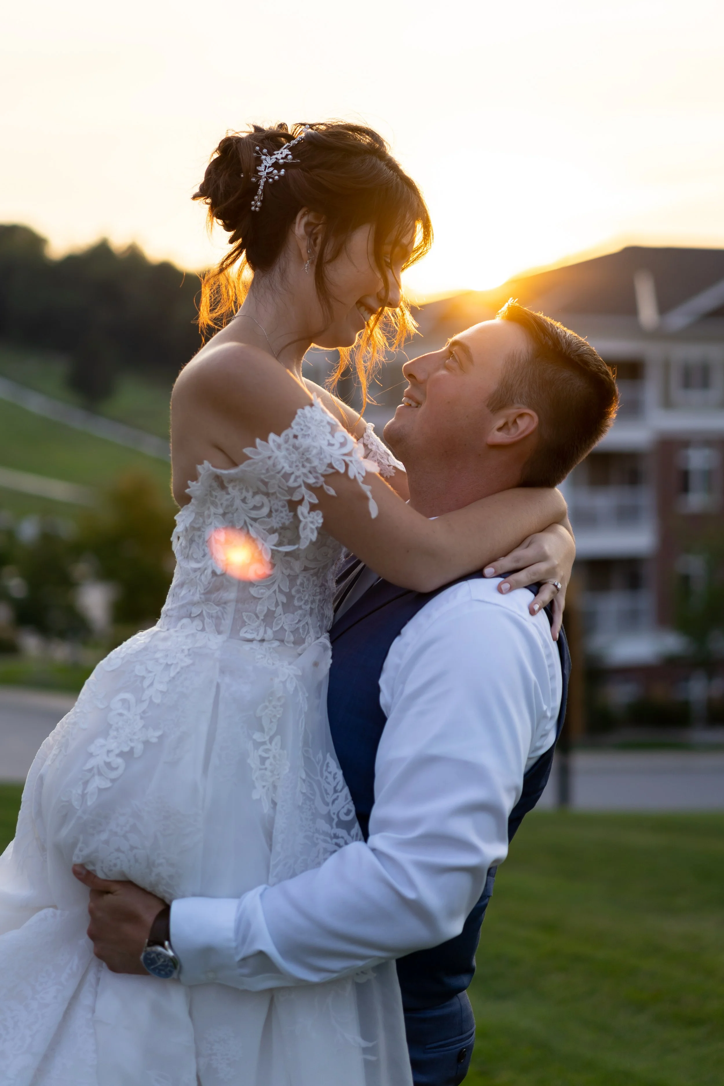 A groom lifting a bride in his arms outdoors during sunset, both smiling and looking at each other, on a grassy area with a house and trees in the background.