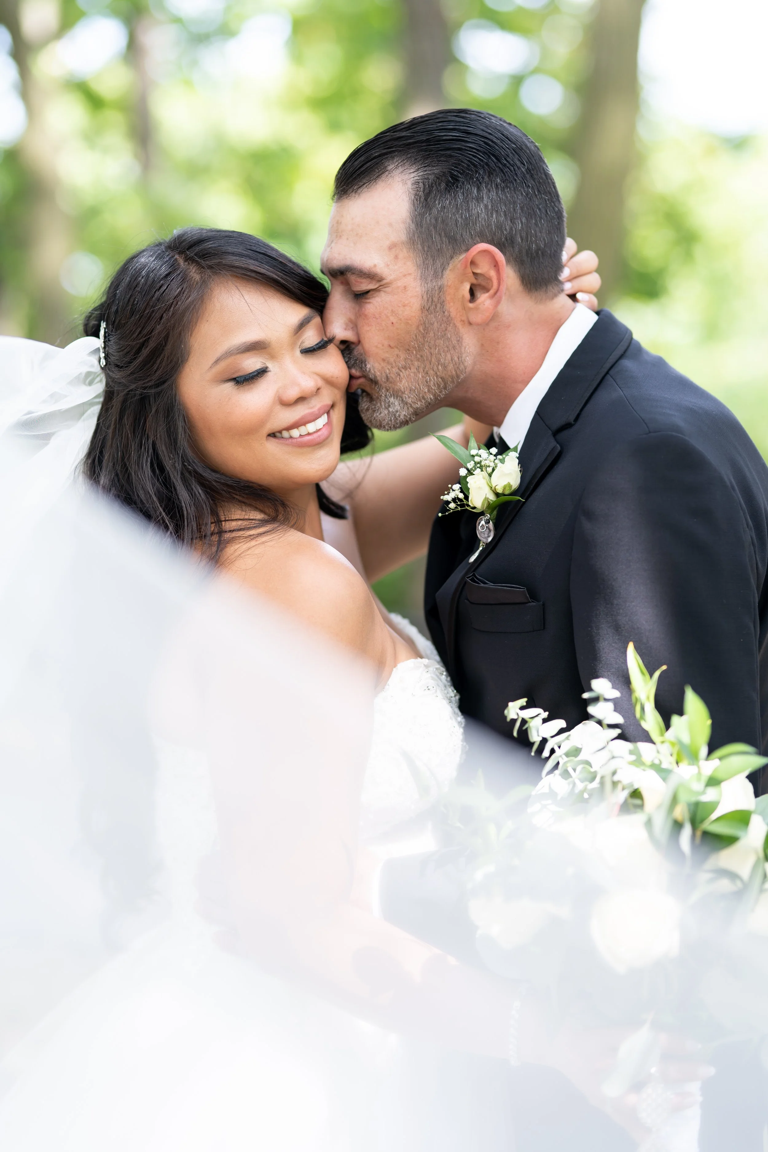 A bride and groom share a kiss outdoors during their wedding, with the bride smiling and the groom wearing a black tuxedo with a white boutonniere, surrounded by greenery.