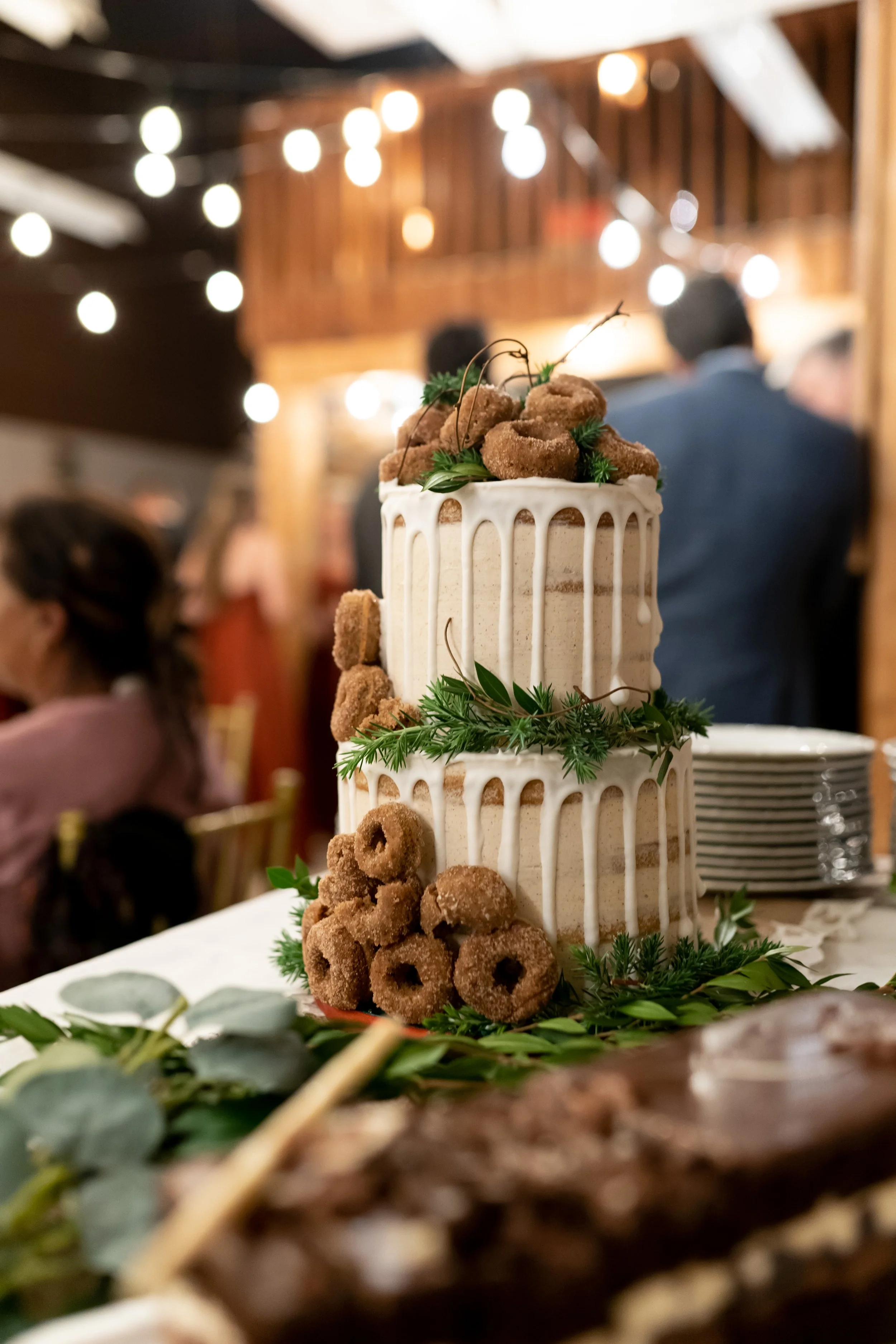 A multi-tiered cake decorated with brown treats and green foliage, in a rustic indoor setting with warm string lights and blurred guests in the background.
