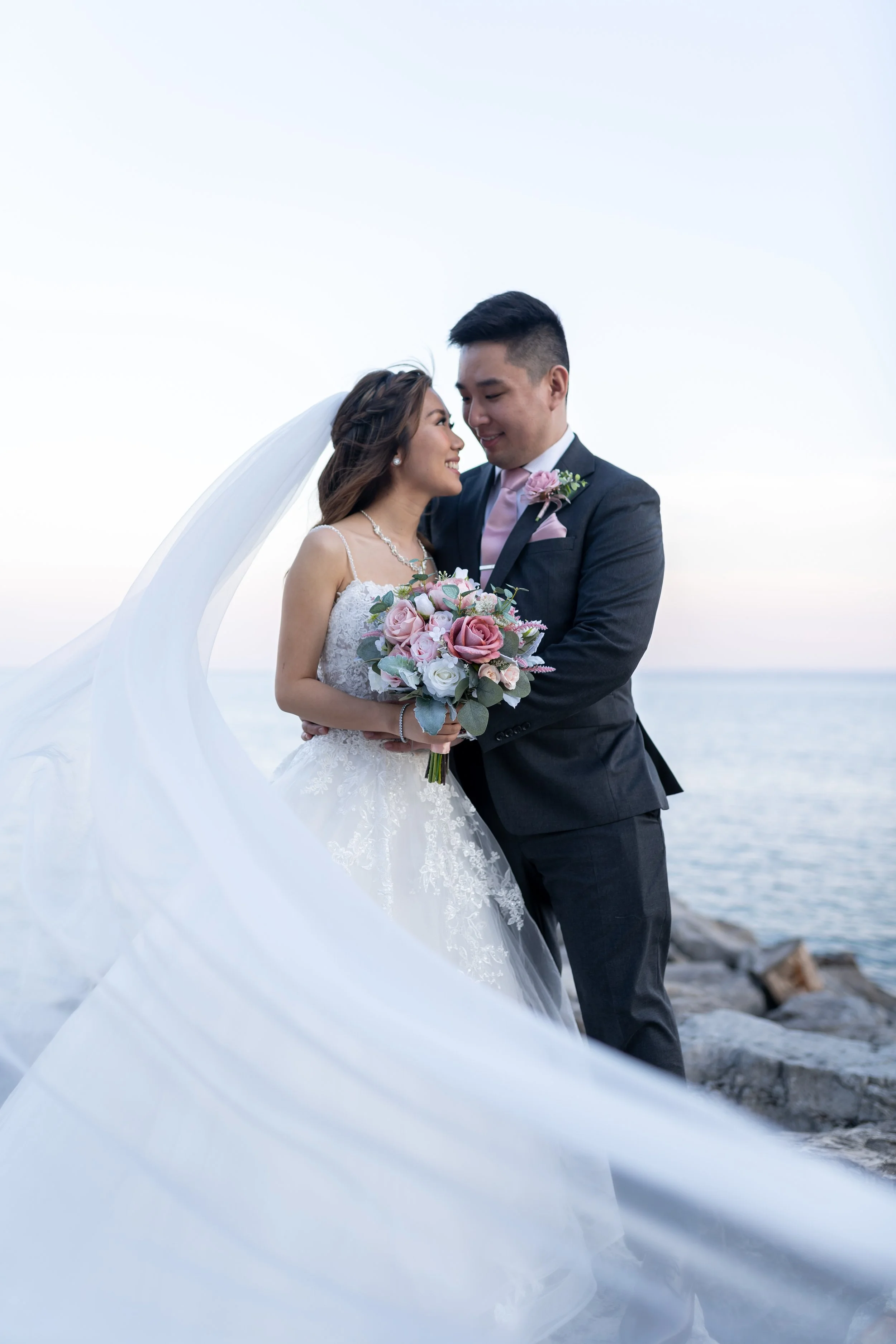 Bride and groom standing by the water, smiling and holding a bouquet of pink and white flowers, with the bride's long veil flowing in the wind, at sunset or early evening.