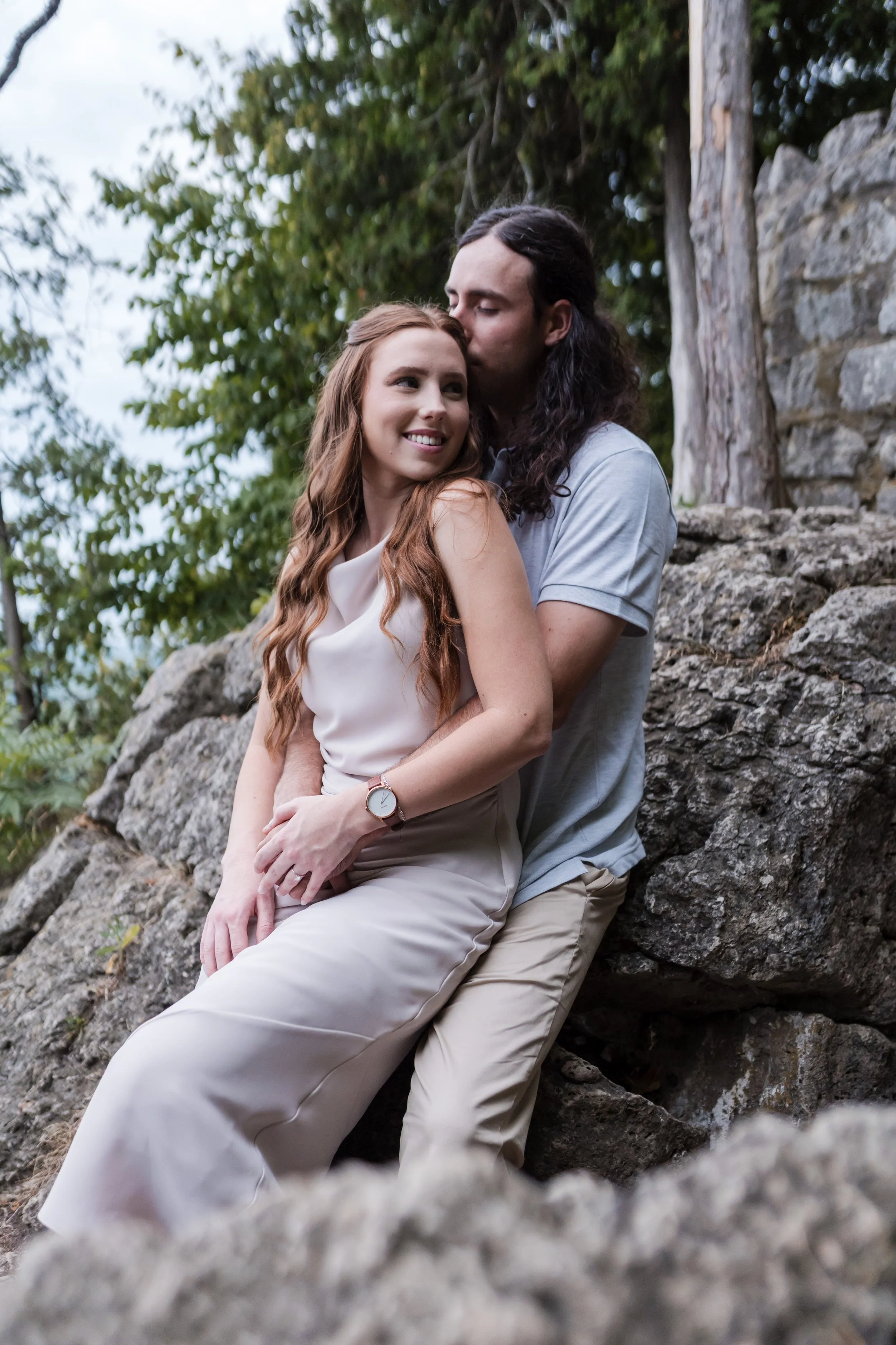 A young couple sitting on rocks outdoors, with the woman smiling and the man kissing her forehead, surrounded by trees.