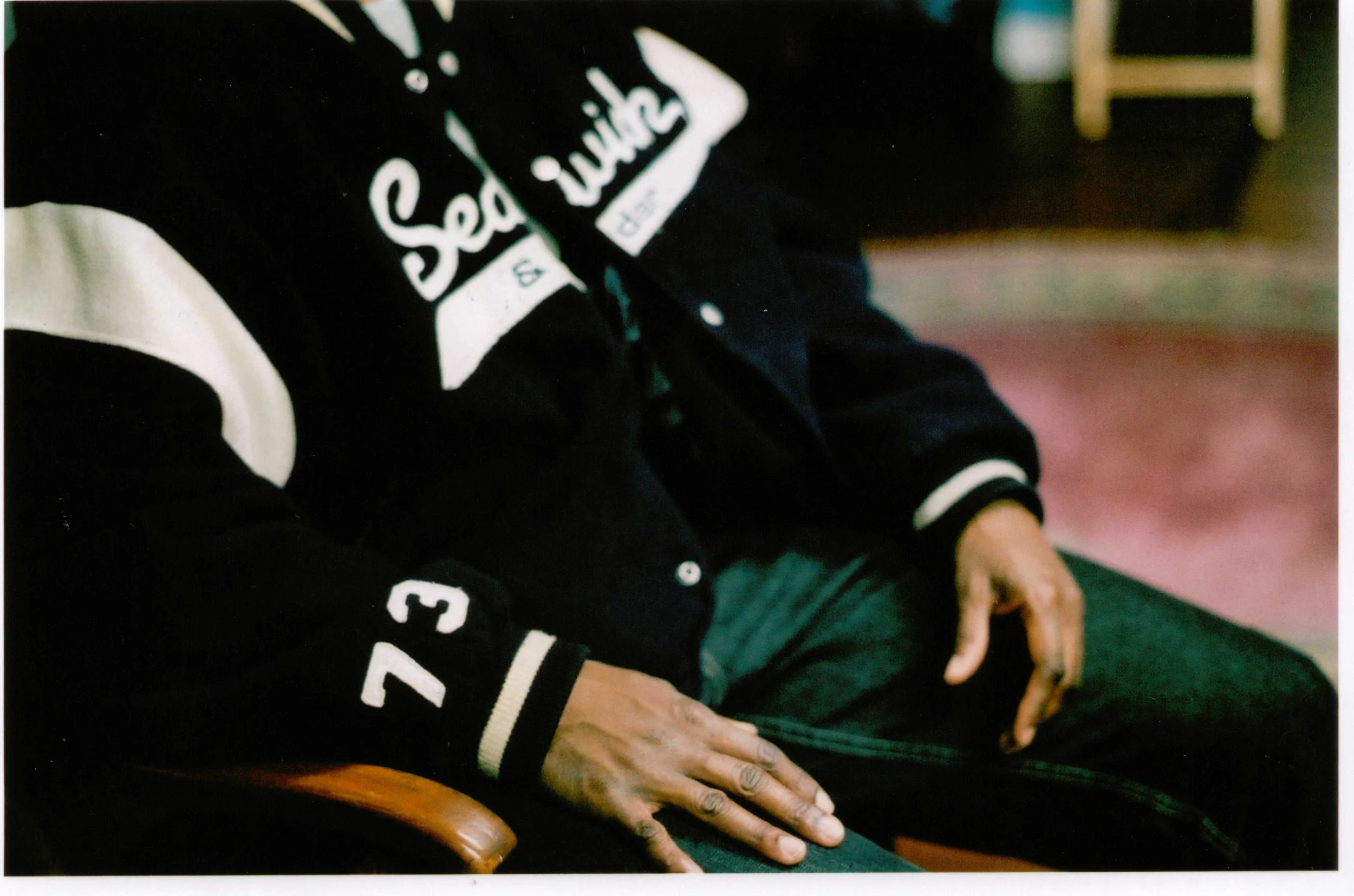 Close-up of a person wearing a black varsity jacket with white lettering and patches, sitting with hands resting on their lap, with another person partially visible beside them.