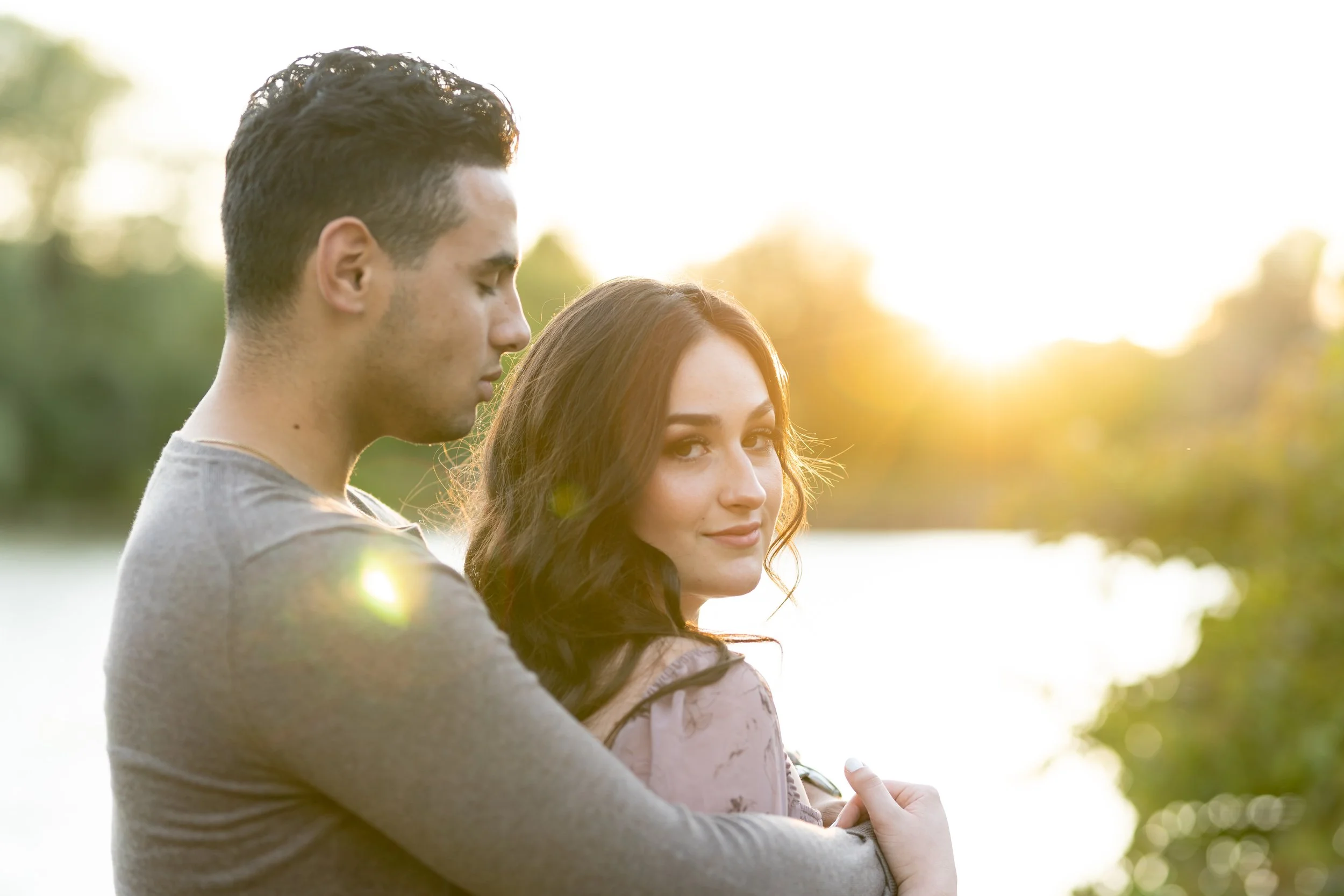 A young man and woman standing close outdoors near a body of water with trees in the background, during sunset, with warm sunlight illuminating them.