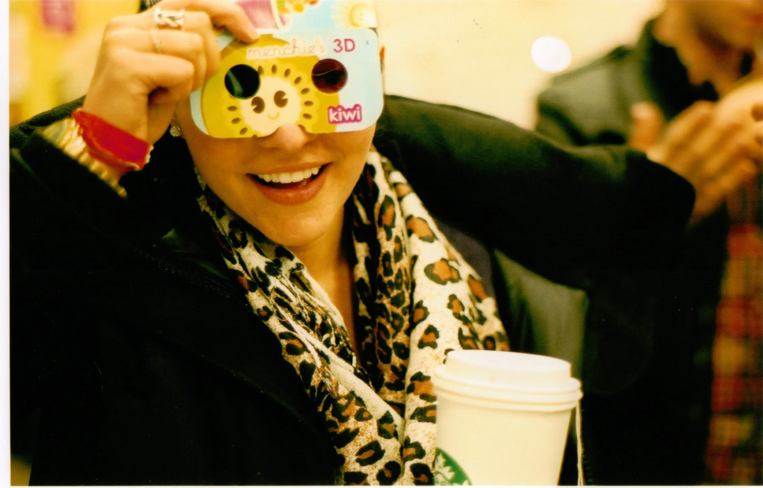 A woman holding a cup with a lid and a straw, wearing a leopard print scarf, sitting indoors, with a friend taking a selfie in the background.