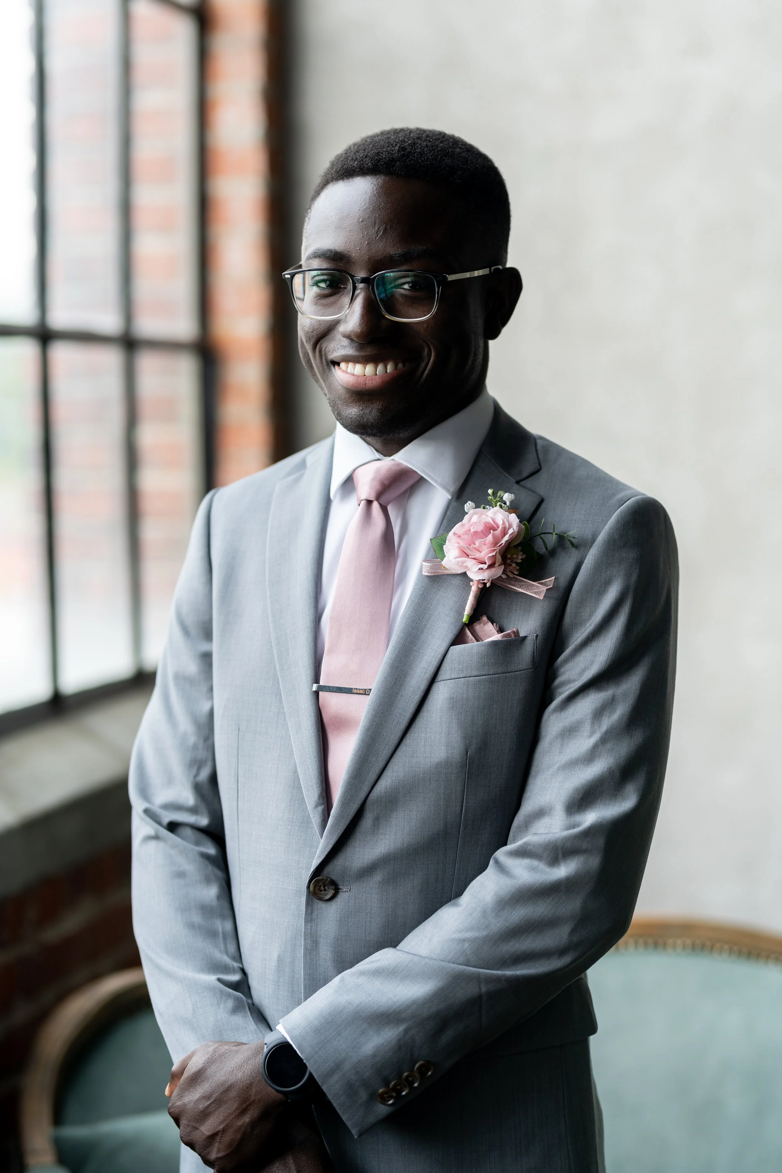 A man in a light gray suit with a pink tie and a pink boutonniere, wearing glasses, smiling and standing near a window with exposed brick and a white wall.