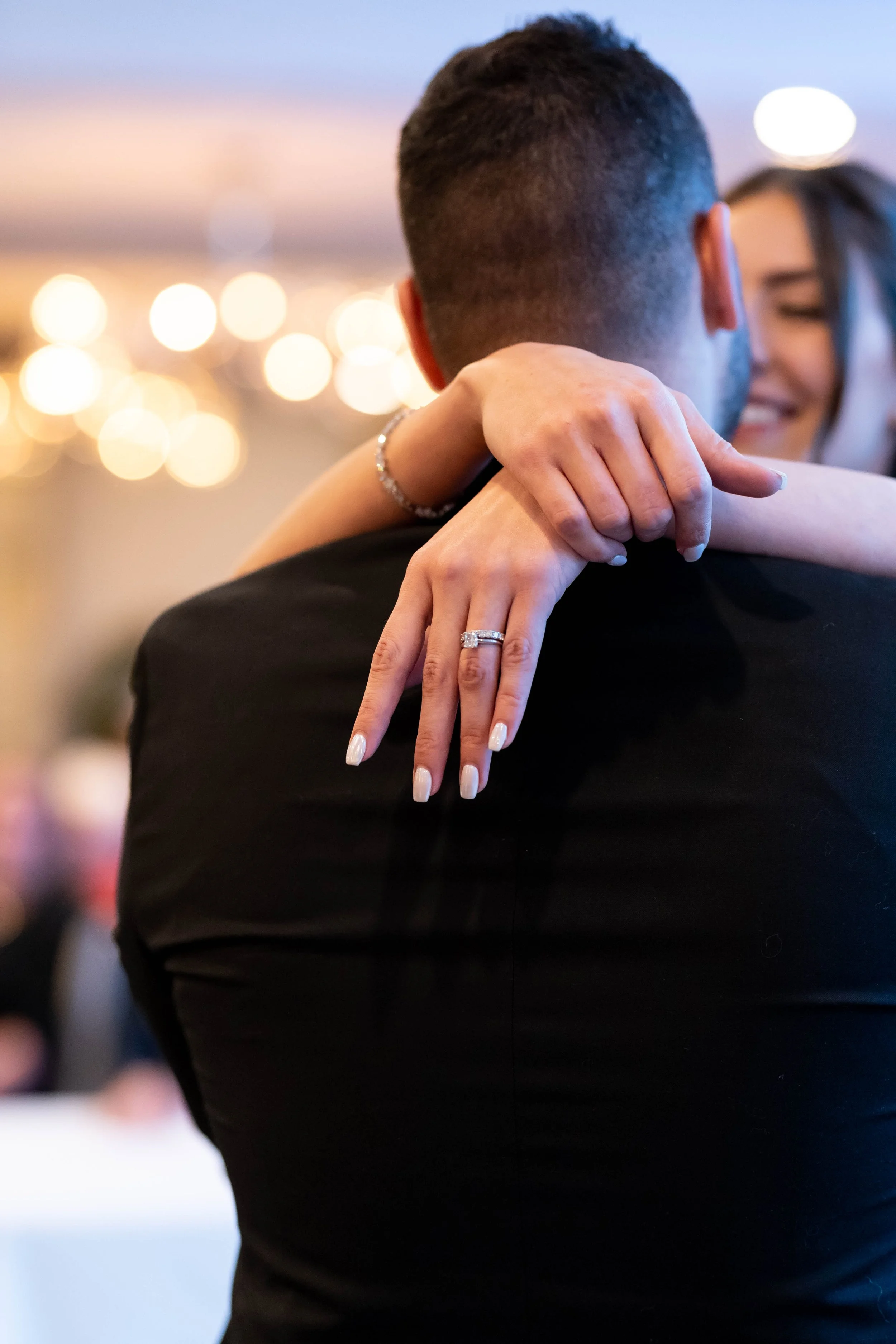 A man and woman dancing closely at an event, with the woman's hand resting on the man's shoulder and the man's back facing the camera. The woman is smiling and wearing a ring and a bracelet. The background has blurred, warm lighting.