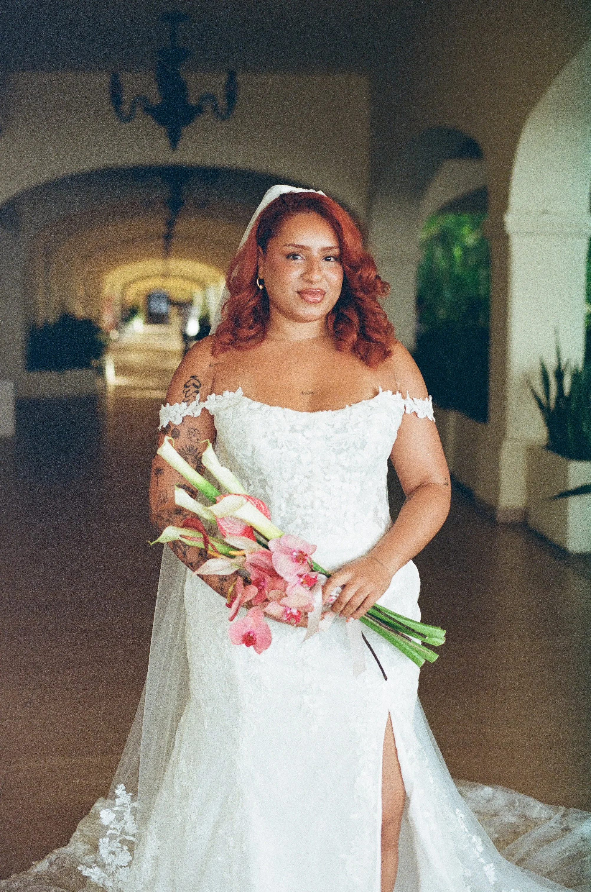 A woman in a white wedding dress holding a bouquet of pink flowers, standing in an indoor hallway with arched ceilings and a chandelier.
