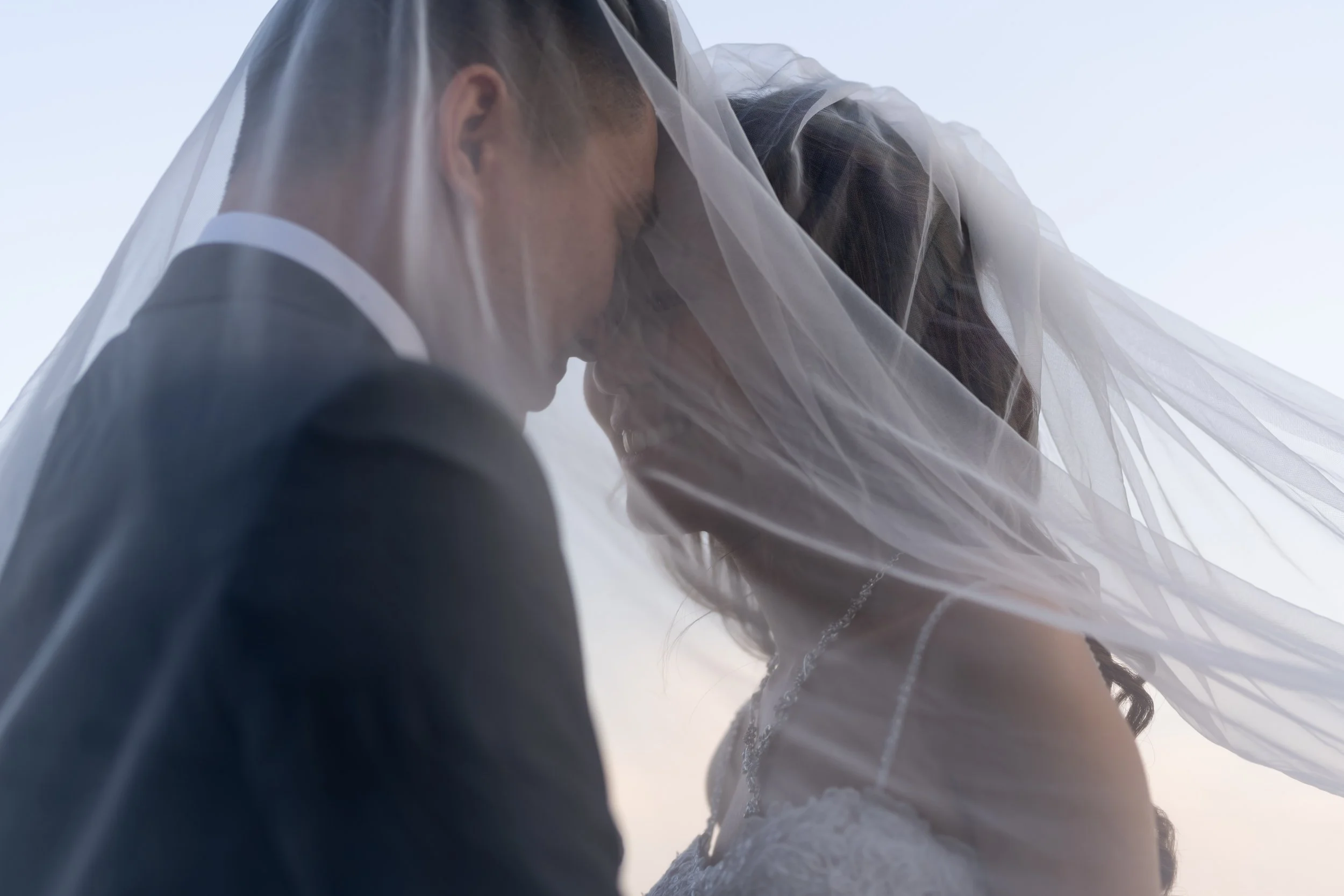 A person in formal attire, possibly a bride, is under a sheer veil outdoors, with a cloudy sky in the background.