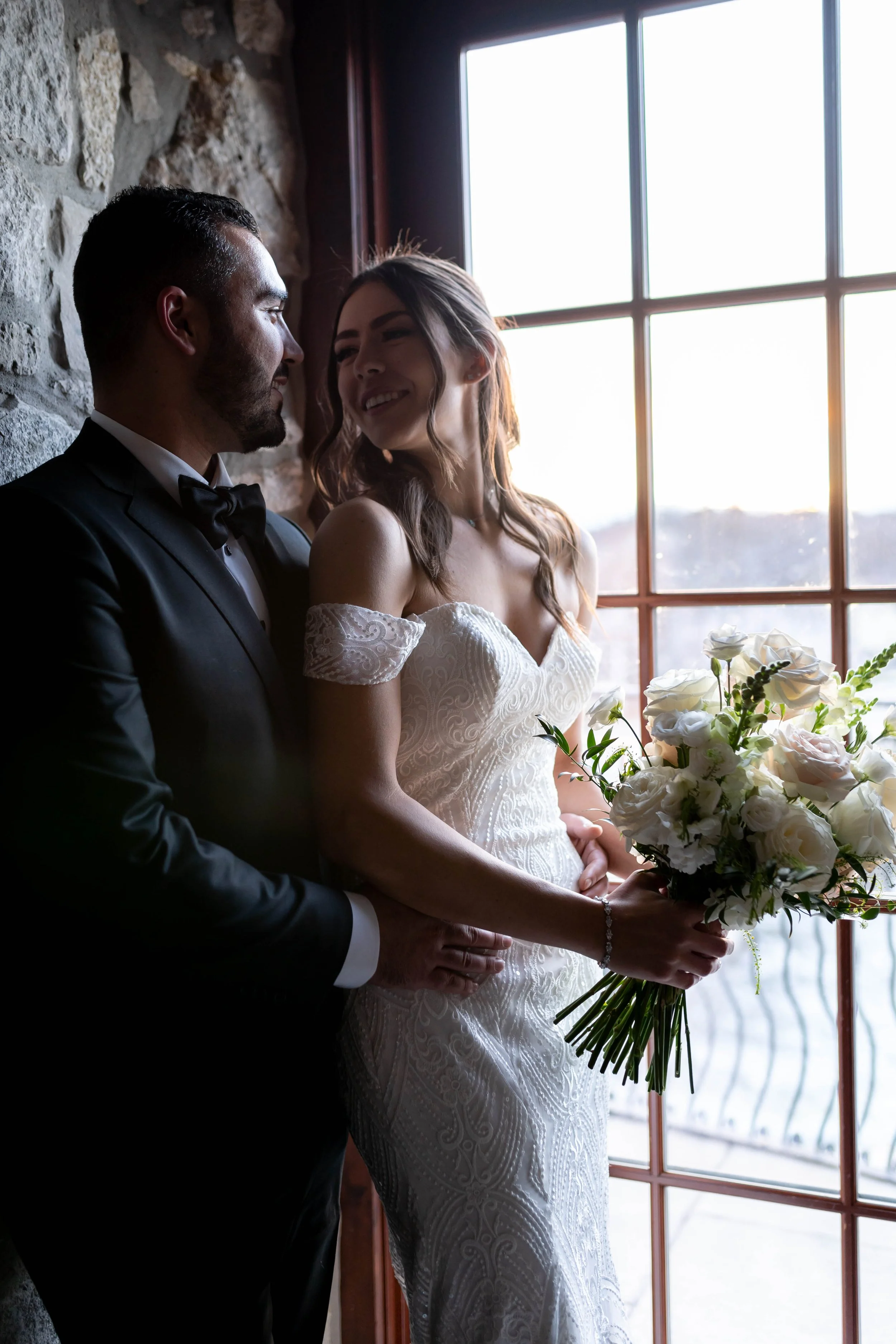 A newlywed couple standing inside by a large window with bright natural light, the groom in a black tuxedo and the bride holding a bouquet of white flowers, both smiling and sharing an intimate moment.