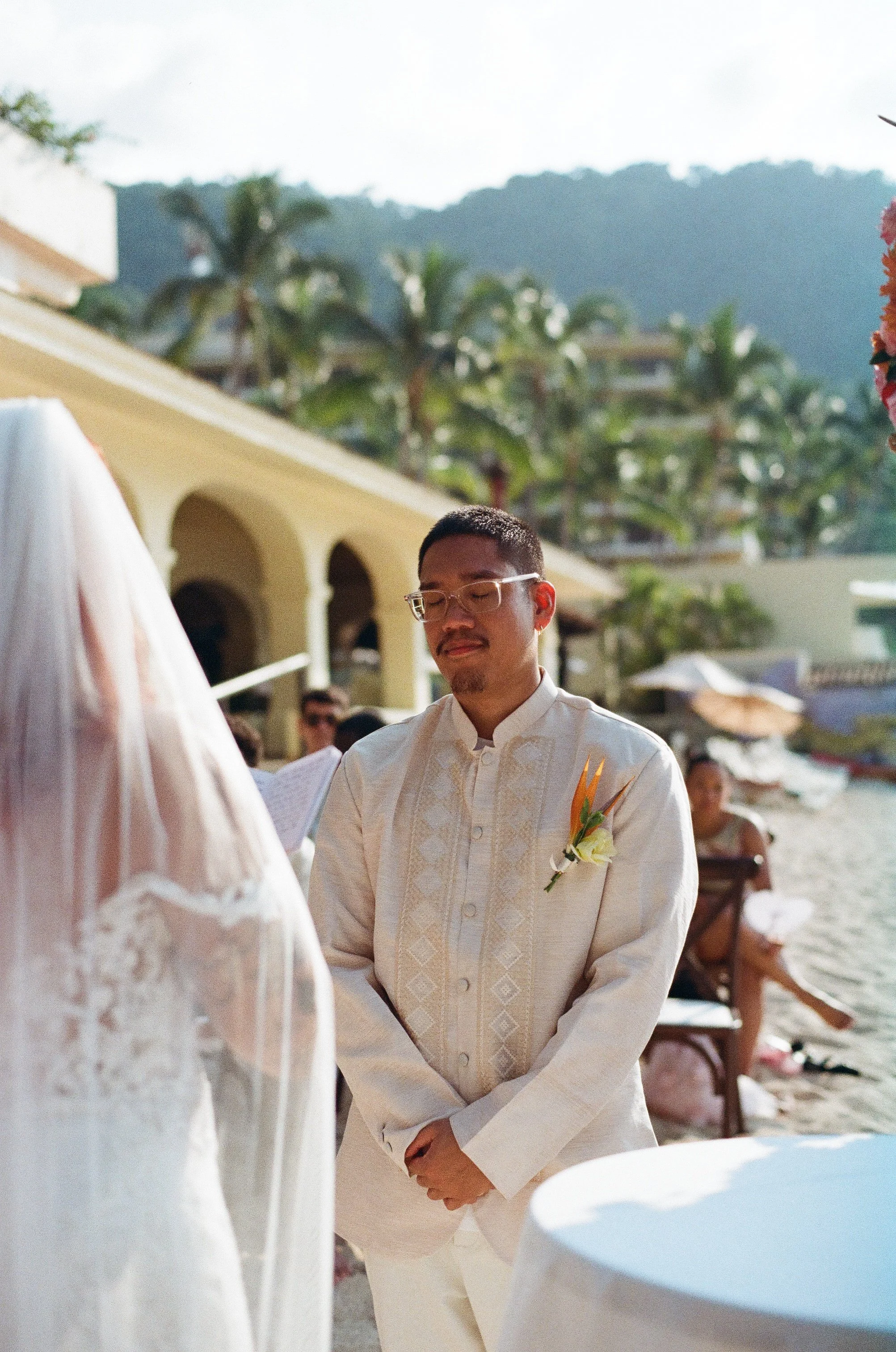 A man dressed in traditional wedding attire with a boutonniere on his chest, during an outdoor wedding ceremony with a beach and mountain backdrop, and people seated under umbrellas.
