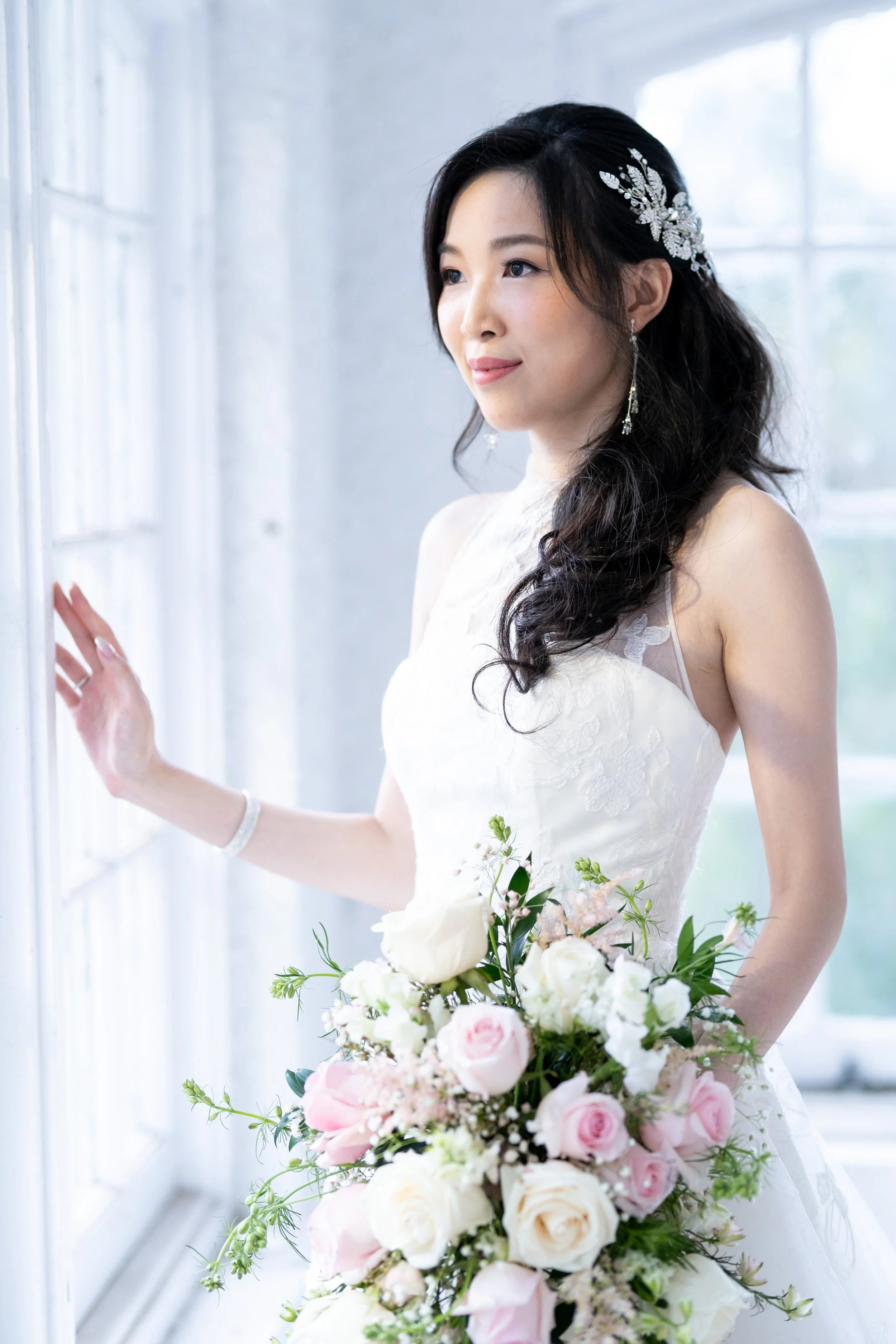 A bride in a white wedding dress holding a large bouquet of pink and white roses, standing near a window with natural light.