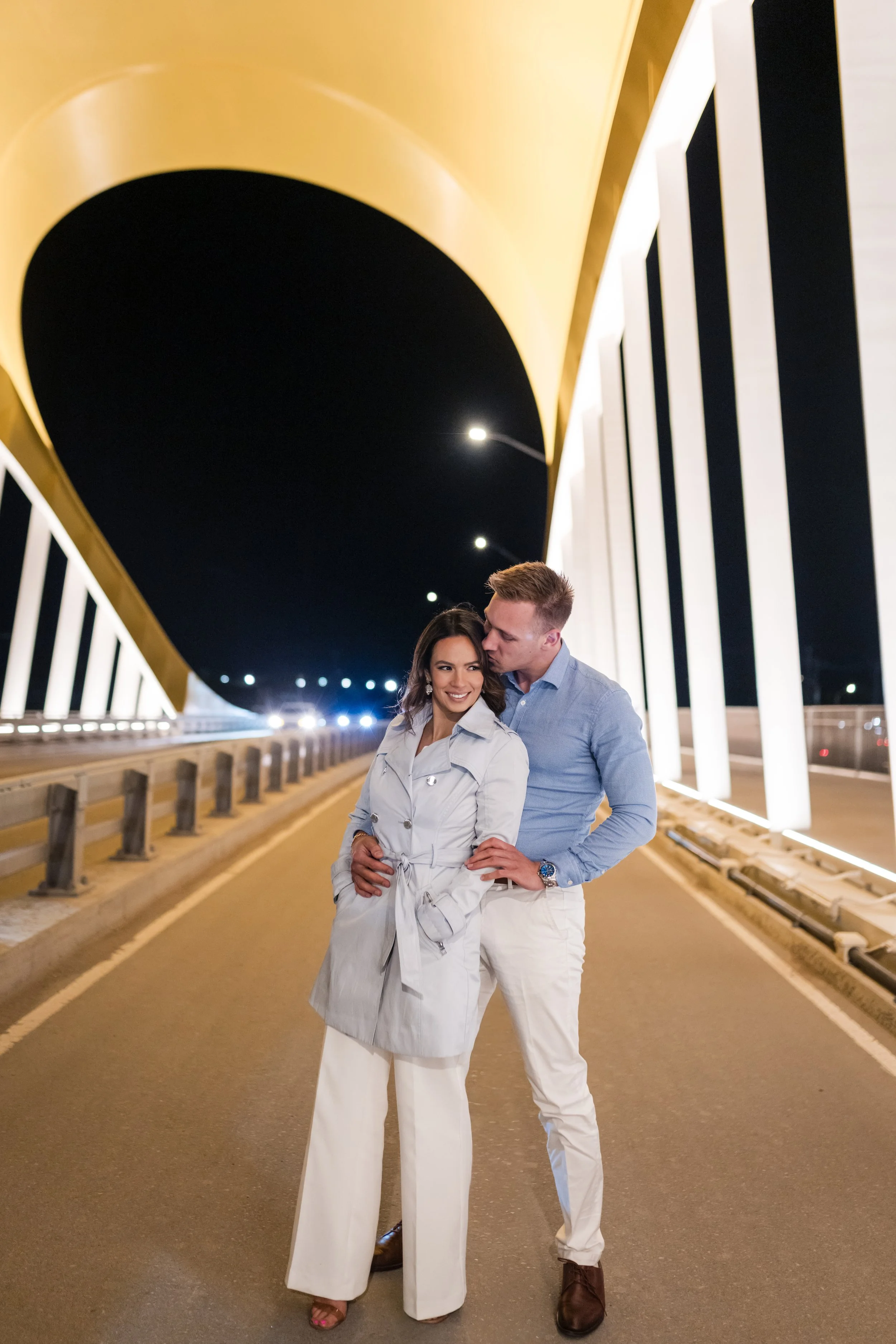 A couple stands close together on a well-lit city street at night, smiling and embracing under a modern, illuminated architectural structure with a black and gold design.