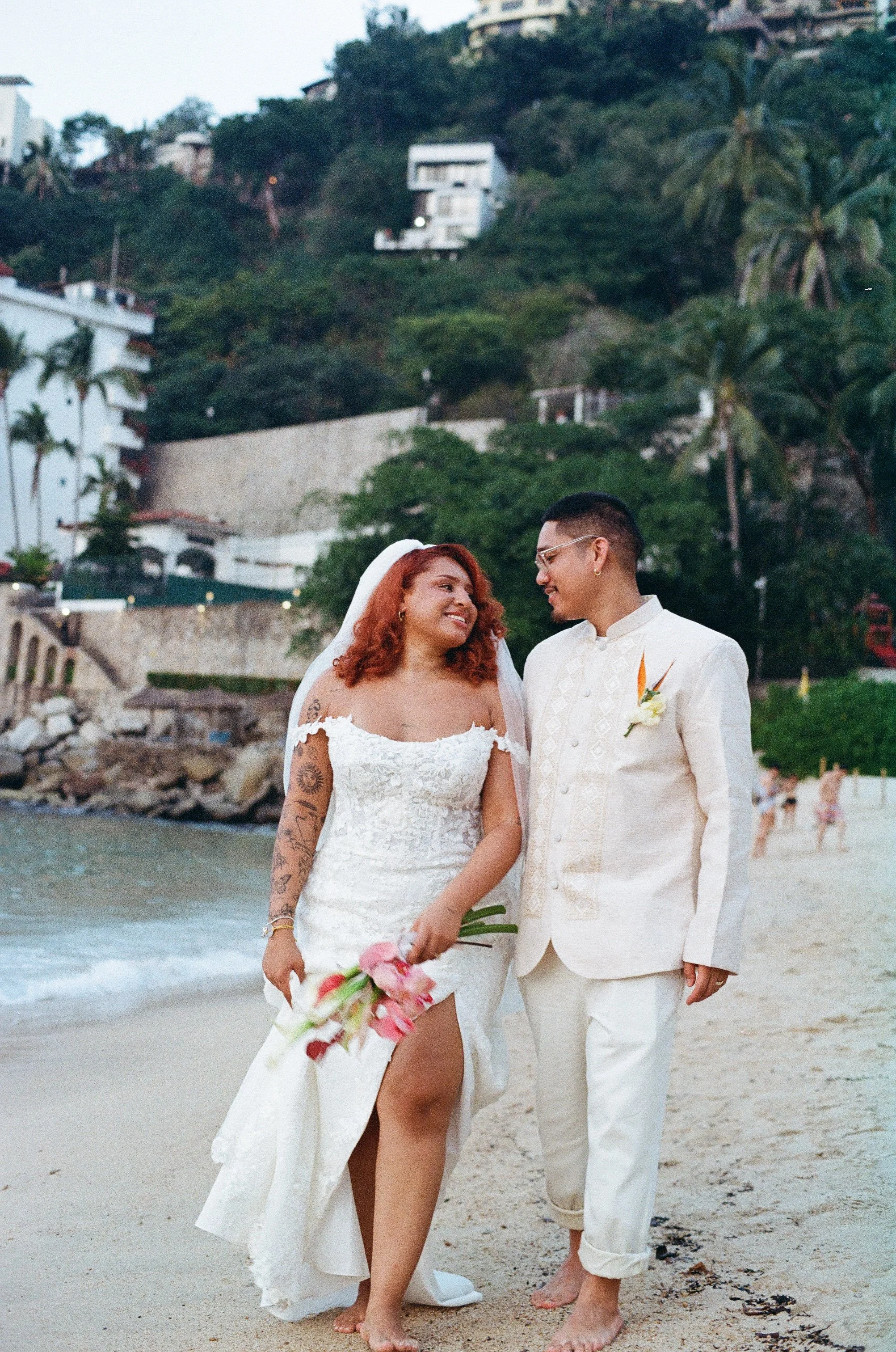 A bride and groom smiling at each other on a beach, holding a bouquet of pink flowers, with palm trees and hillside houses in the background.