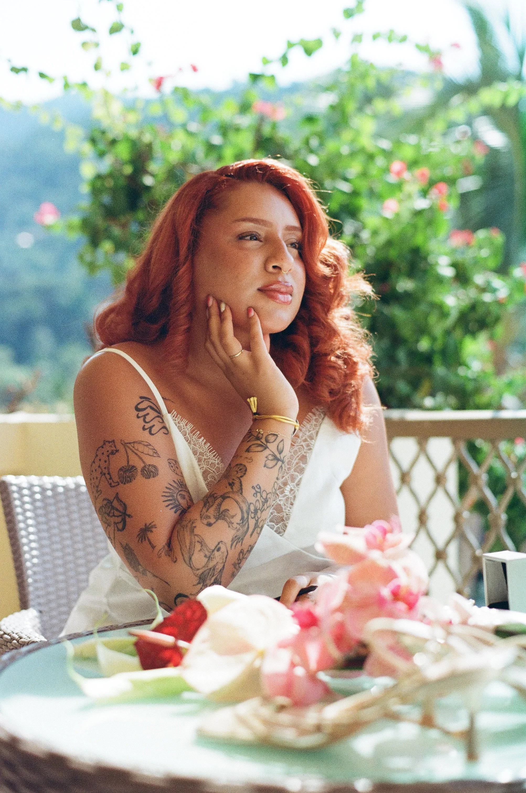 A woman with red hair and tattoos on her arms sits at a table with desserts, in an outdoor setting with greenery in the background.