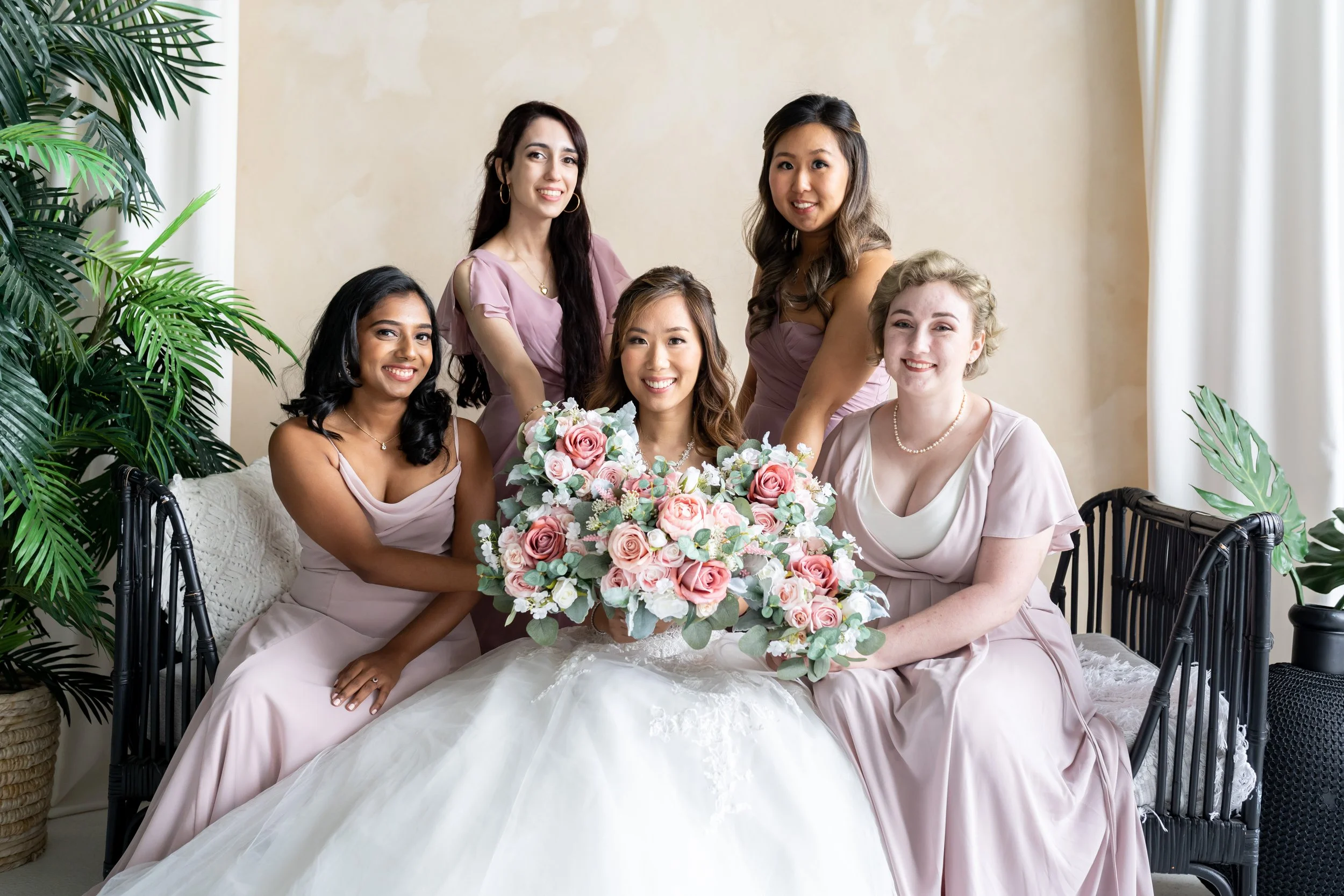 Group of six women dressed in matching light pink dresses, sitting and standing around a table with a large bouquet of pink and white roses, in a room with beige walls and green plants.