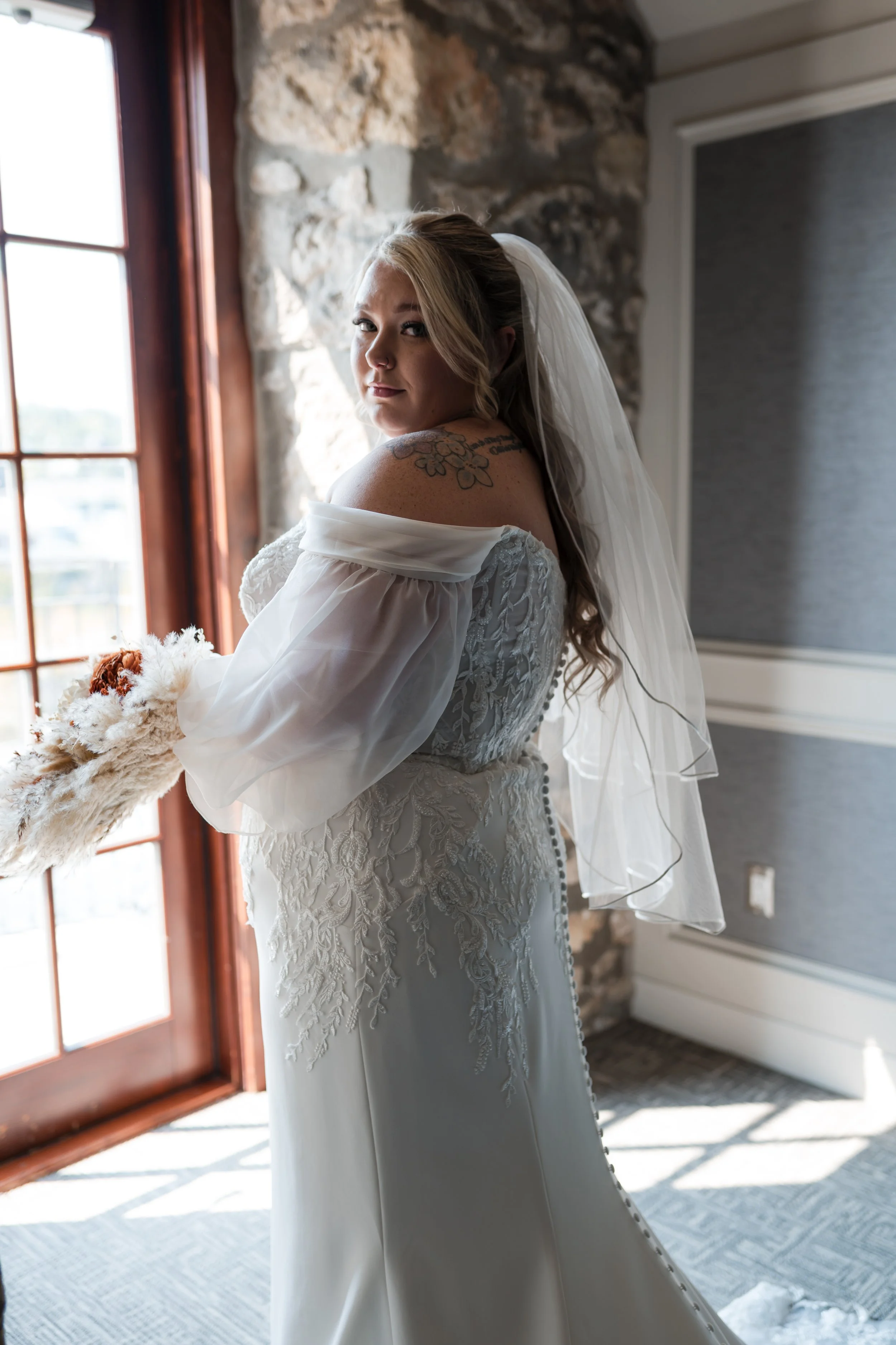 Bride in wedding dress with veil standing by a window with natural light.