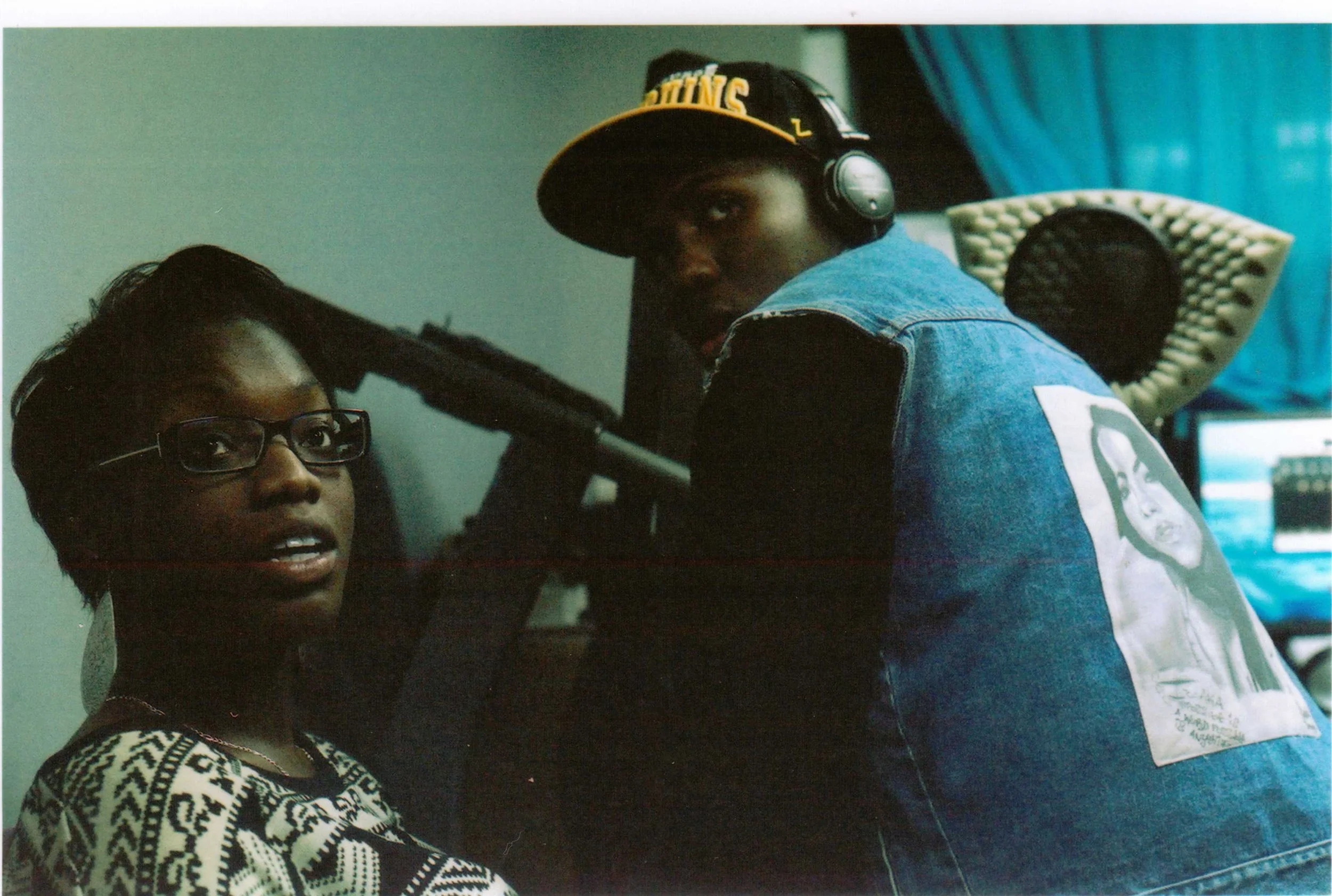 A woman with glasses and a patterned sweater is holding a microphone and speaking, with a young man wearing a baseball cap and headphones leaning in to listen.