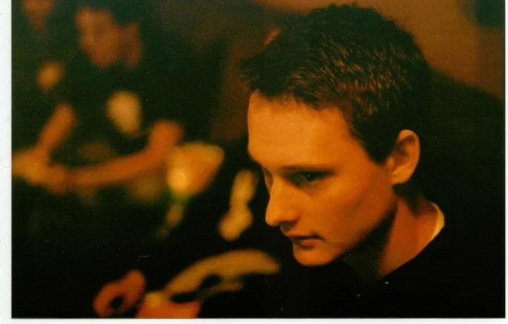 A close-up of a young man with short dark hair, looking down in a dimly lit room with blurred figures in the background.