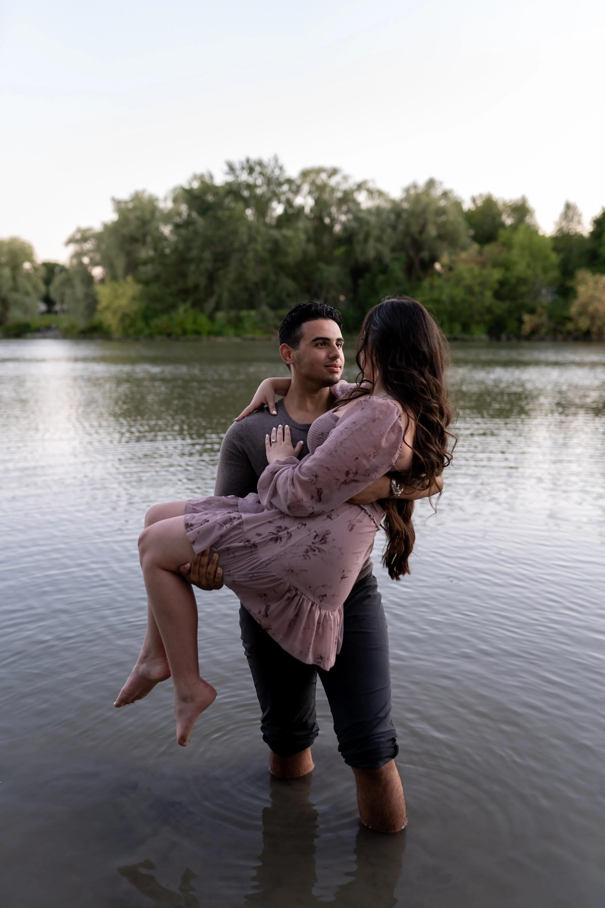 A man in black pants is standing in water up to his knees, holding a woman in pink dress with floral patterns, who is sitting on his arms and legs. They are near a body of water with trees in the background.