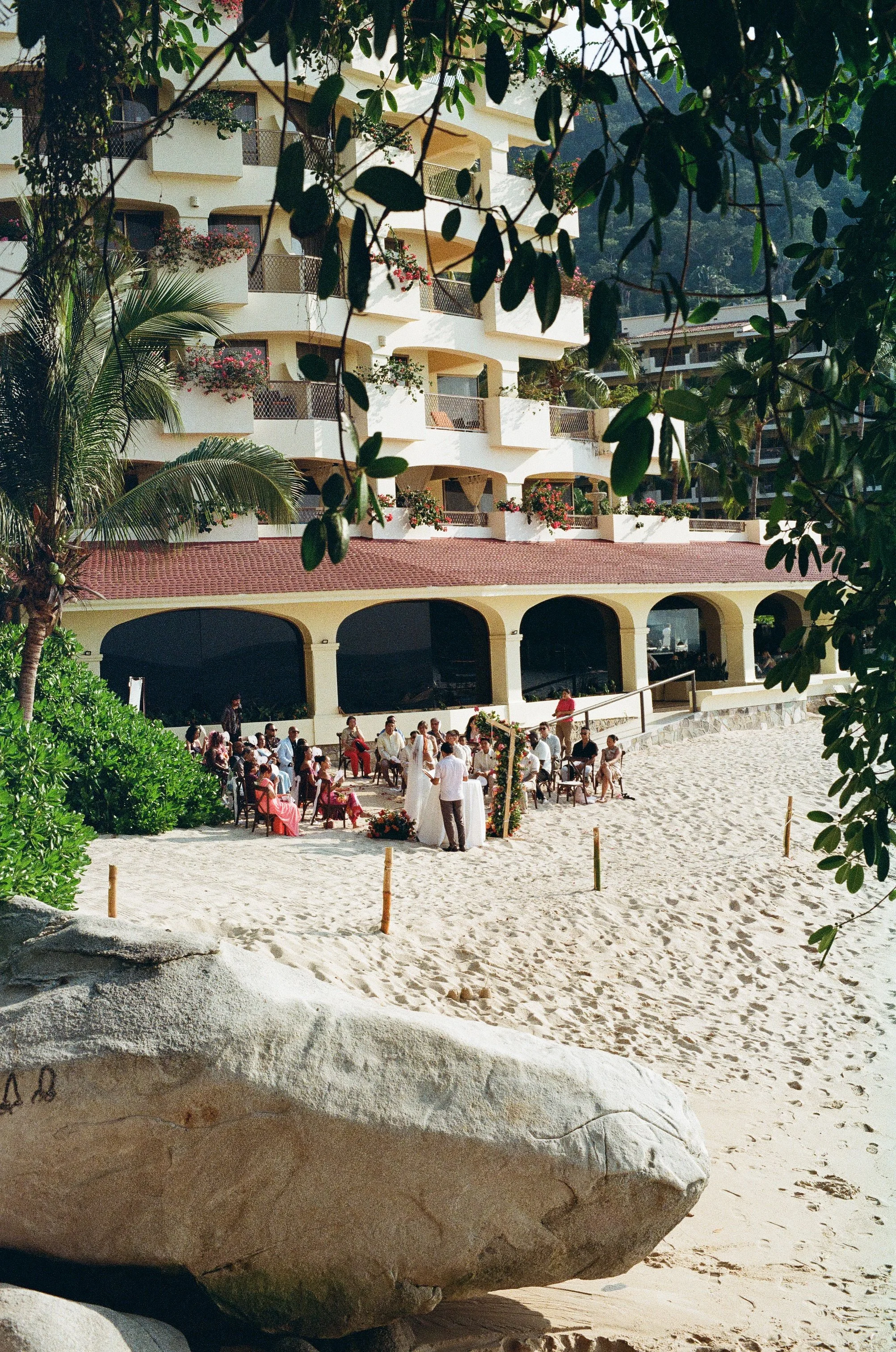 A beach scene with a large rock in the foreground, a sandy area with people sitting and standing, and a multi-story hotel or apartment building overlooking the beach. There are trees and plants around the area.
