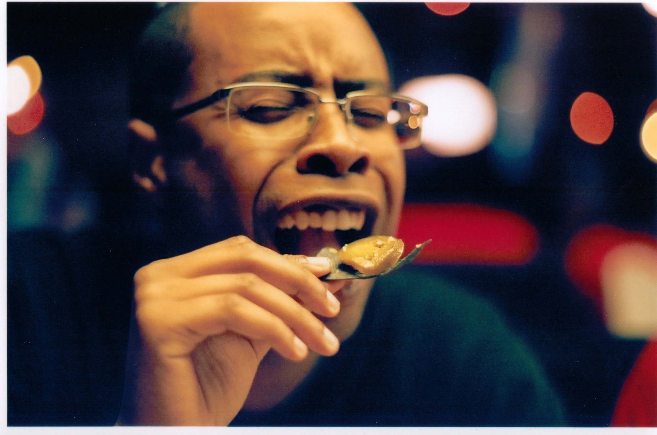 A woman with glasses is smiling with her eyes closed while eating food with a spoon, with blurred colorful lights in the background.