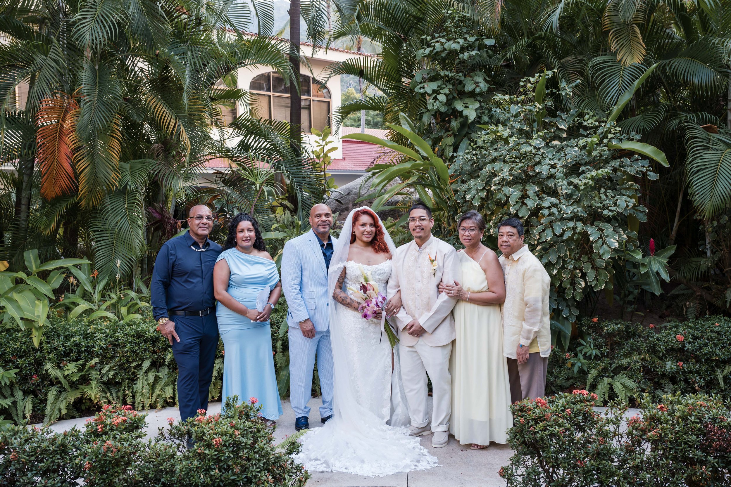 A group of eight diverse people dressed in formal clothing, including a bride in a white wedding dress and a veiled bridegroom in a cream-colored suit, standing outdoors in front of lush tropical greenery puerto vallarta mexico
