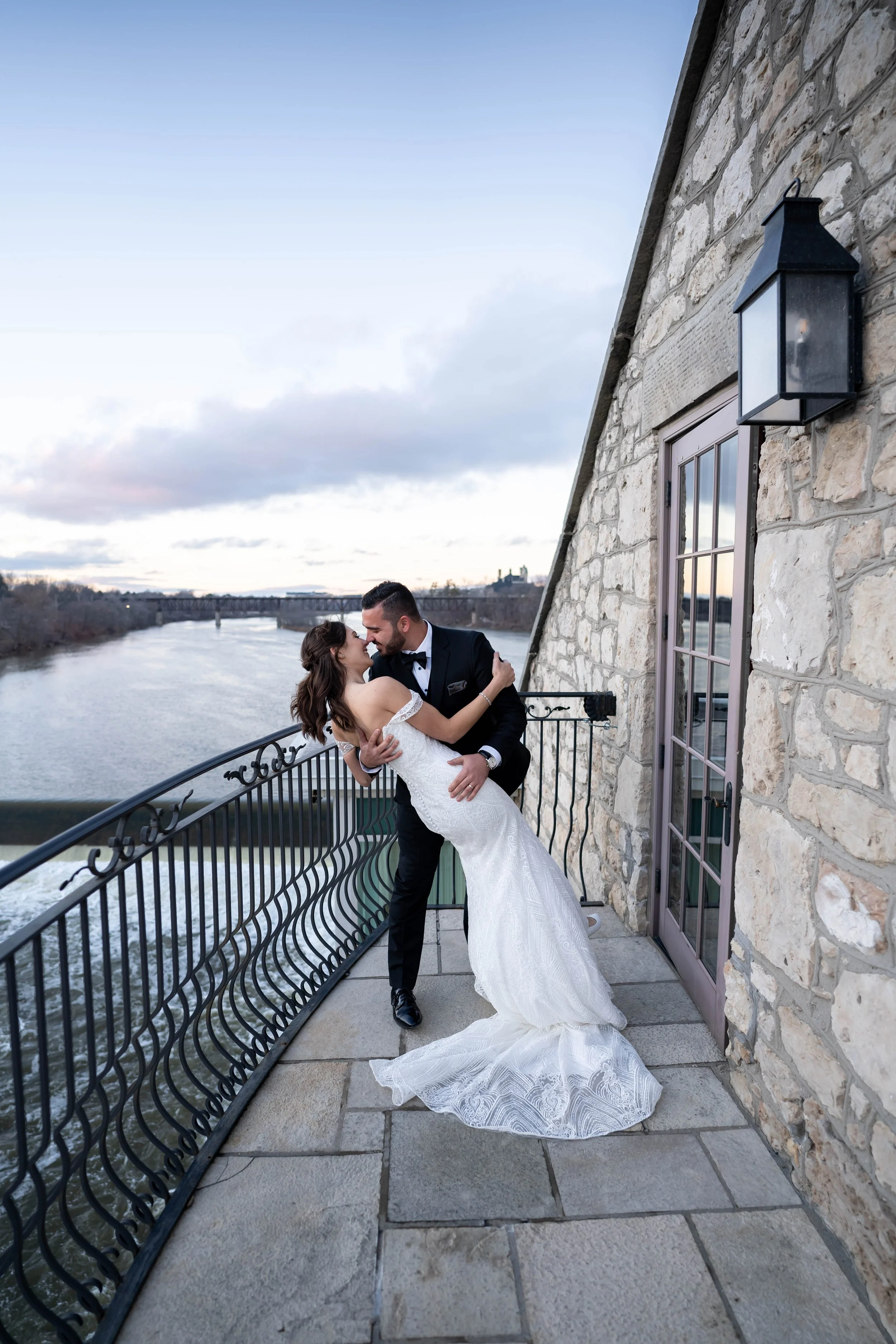 A bride and groom dancing on a balcony overlooking a river, with a stone building wall on the right, a window, and a lantern on the wall. The bride is wearing a white wedding gown, and the groom is in a black tuxedo.