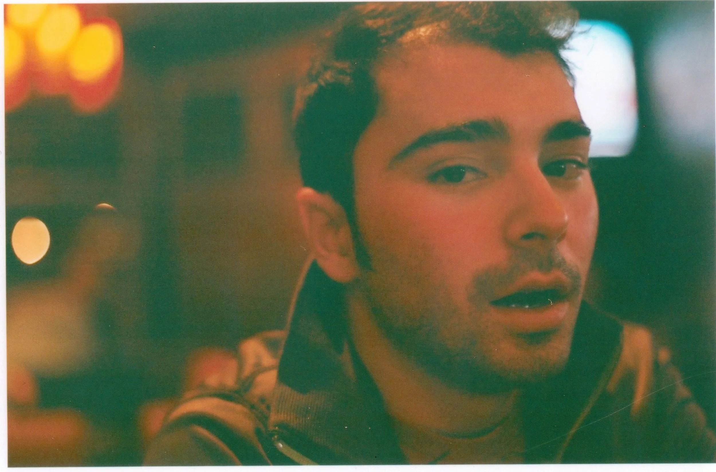 Close-up of a young man with short dark hair and a beard, looking to the side, with background lights creating a bokeh effect.