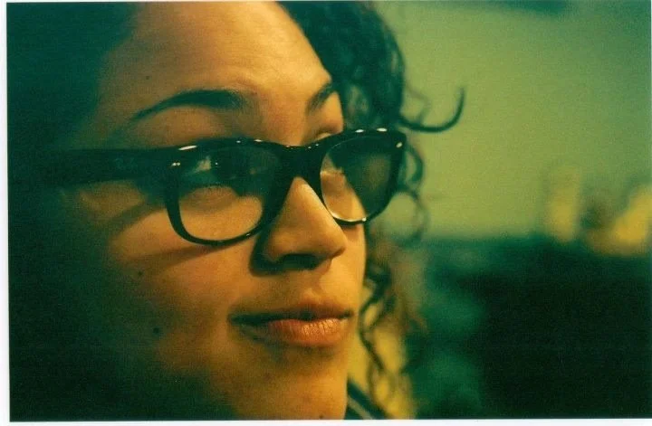 Close-up of a woman wearing black glasses with curly hair and slightly pursed lips.