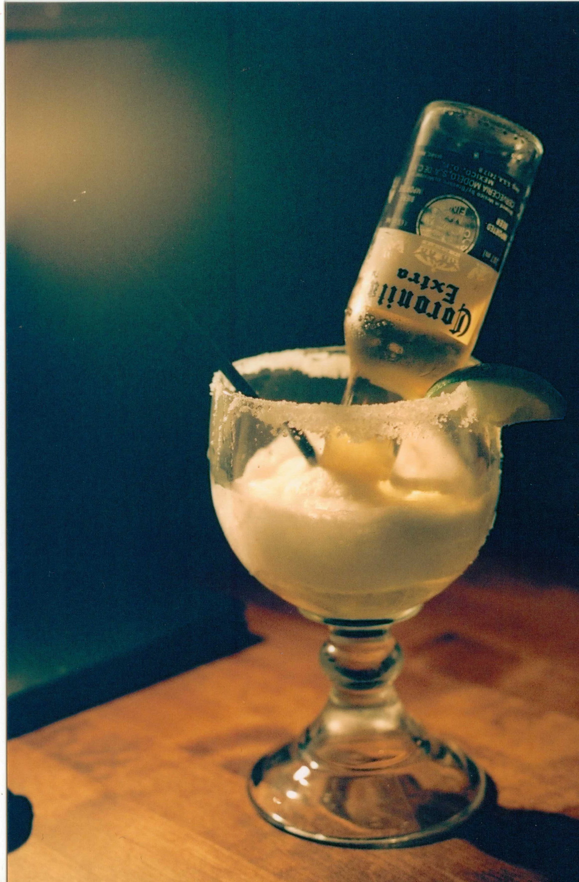 A glass of margarita cocktail with a salt rim, garnished with a lime wedge, and a bottle of Corona beer being poured into the drink.