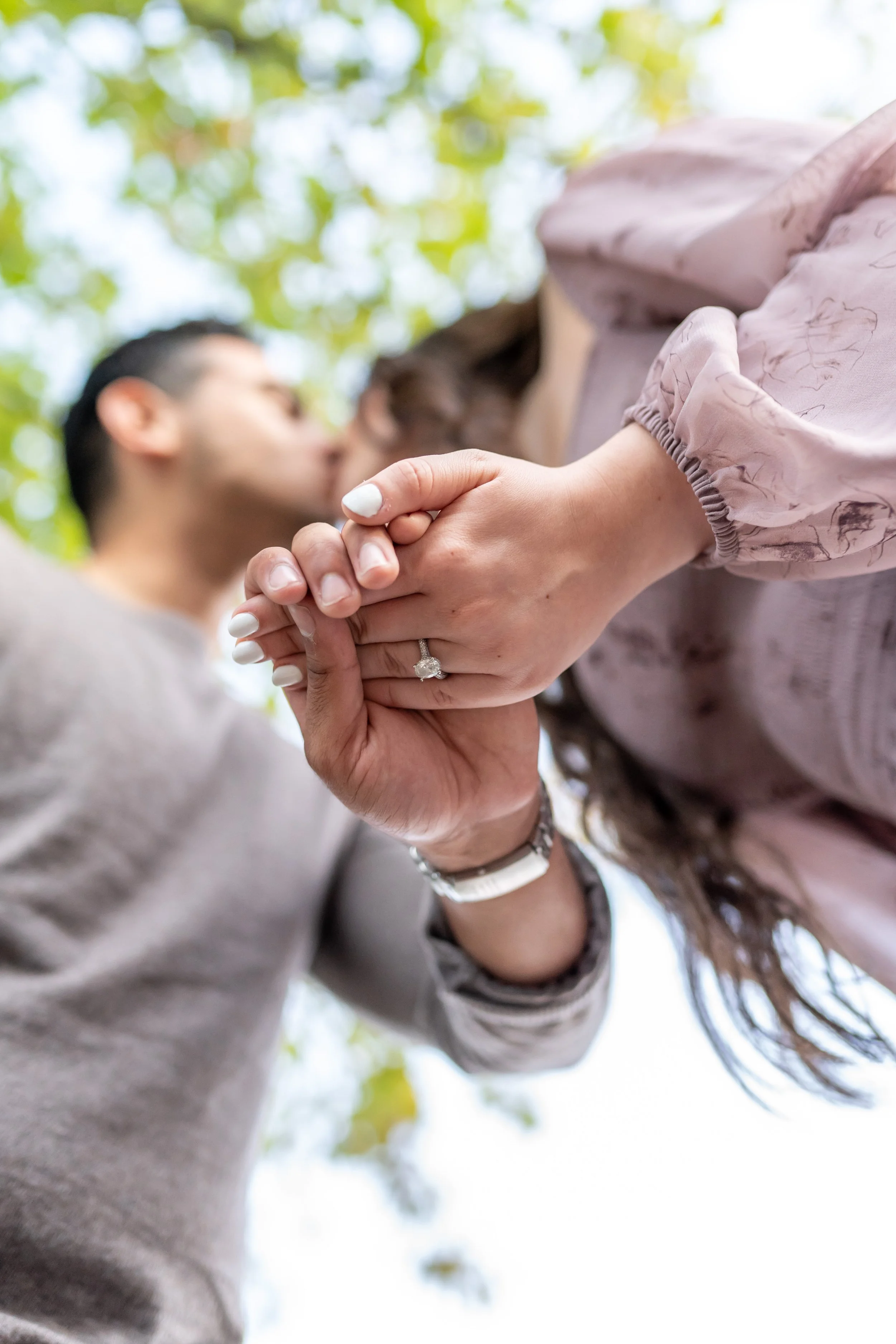 A couple kissing outdoors, holding hands with wedding rings, under a canopy of green leaves.