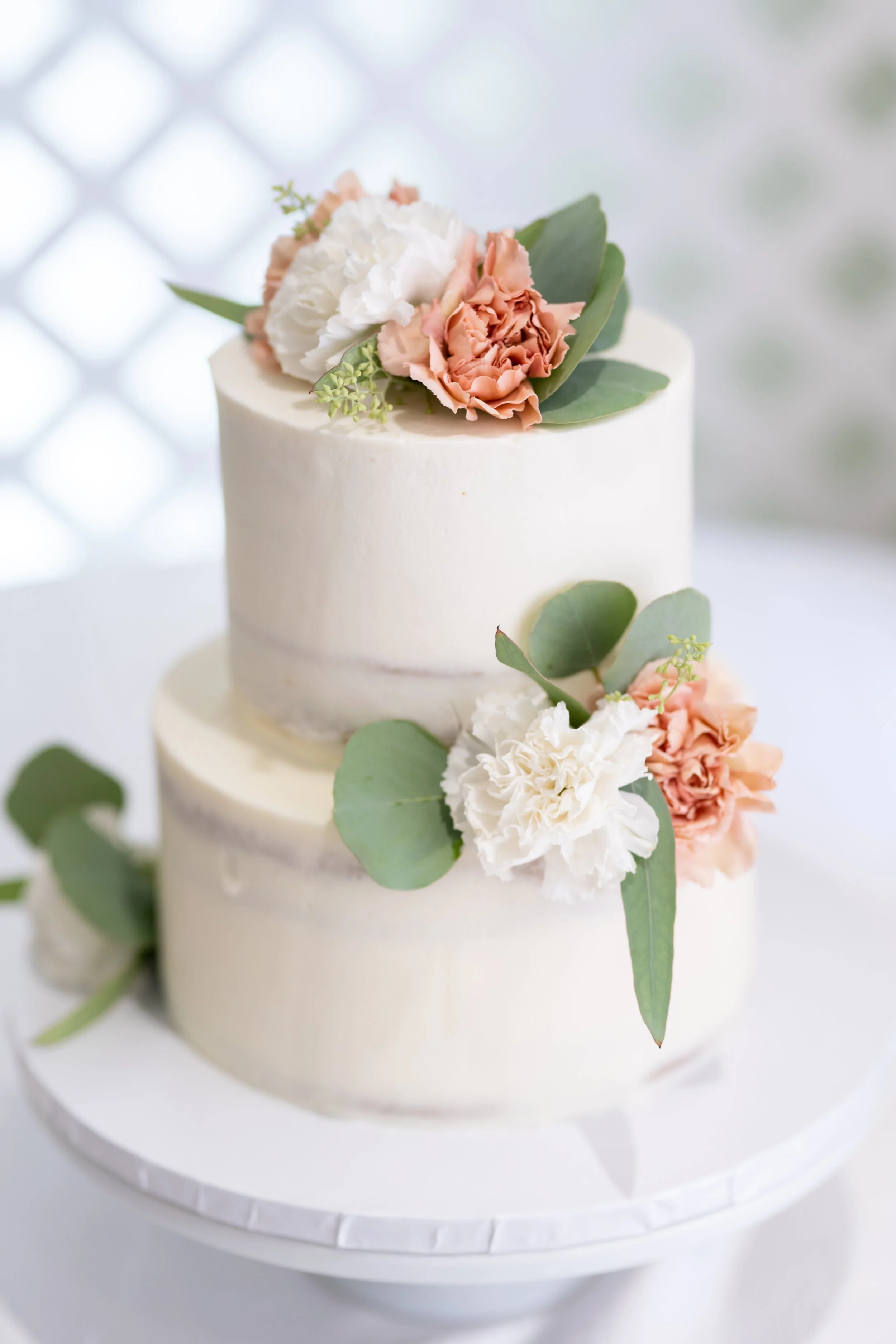 A two-tier white wedding cake decorated with peach and white carnations, green leaves, and small sprigs of greenery, on a white cake stand.