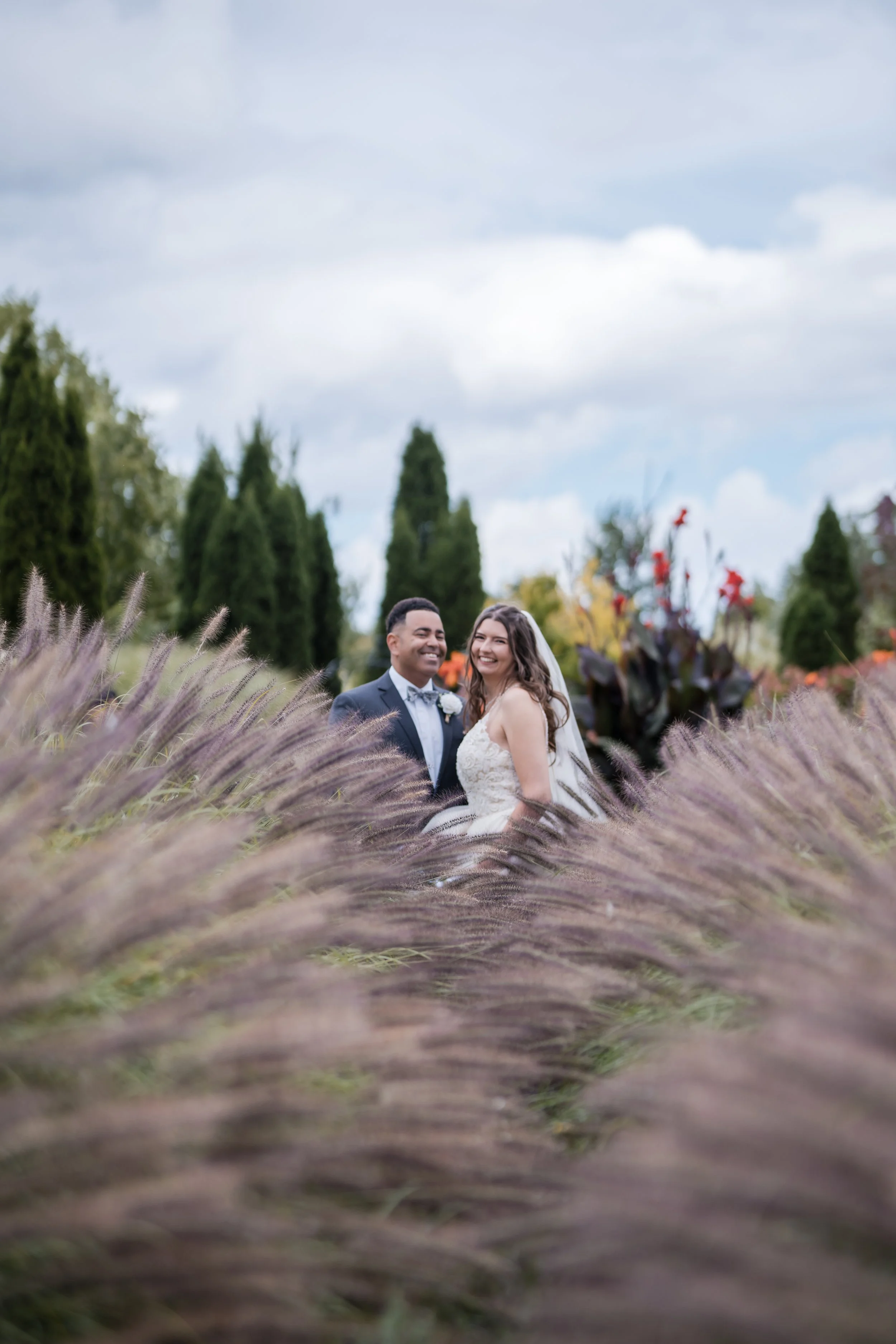 A smiling couple in wedding attire, standing outdoors among tall, purple flowers and lush green trees under a partly cloudy sky.