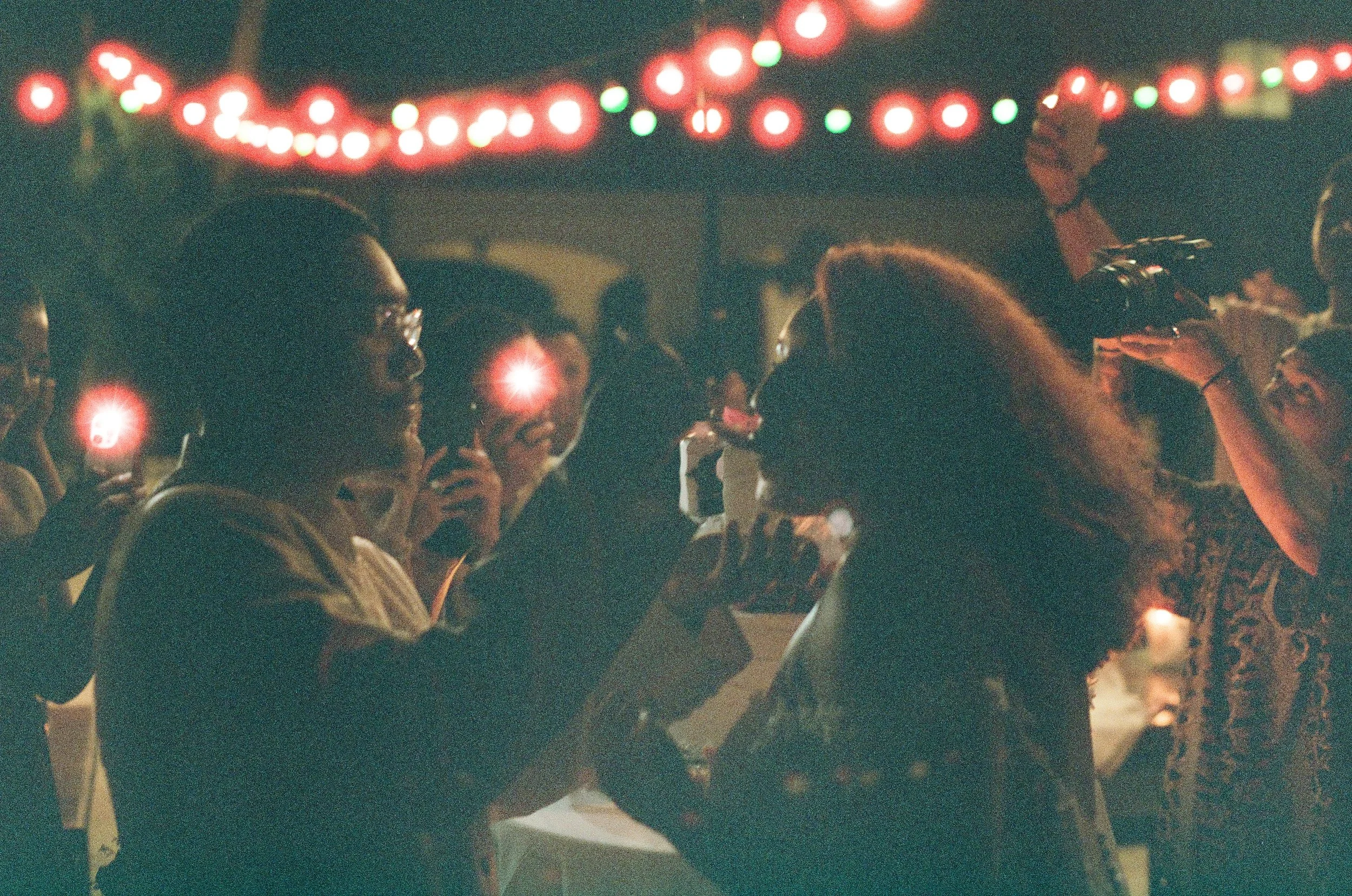 People holding lit candles under festive string lights at night.