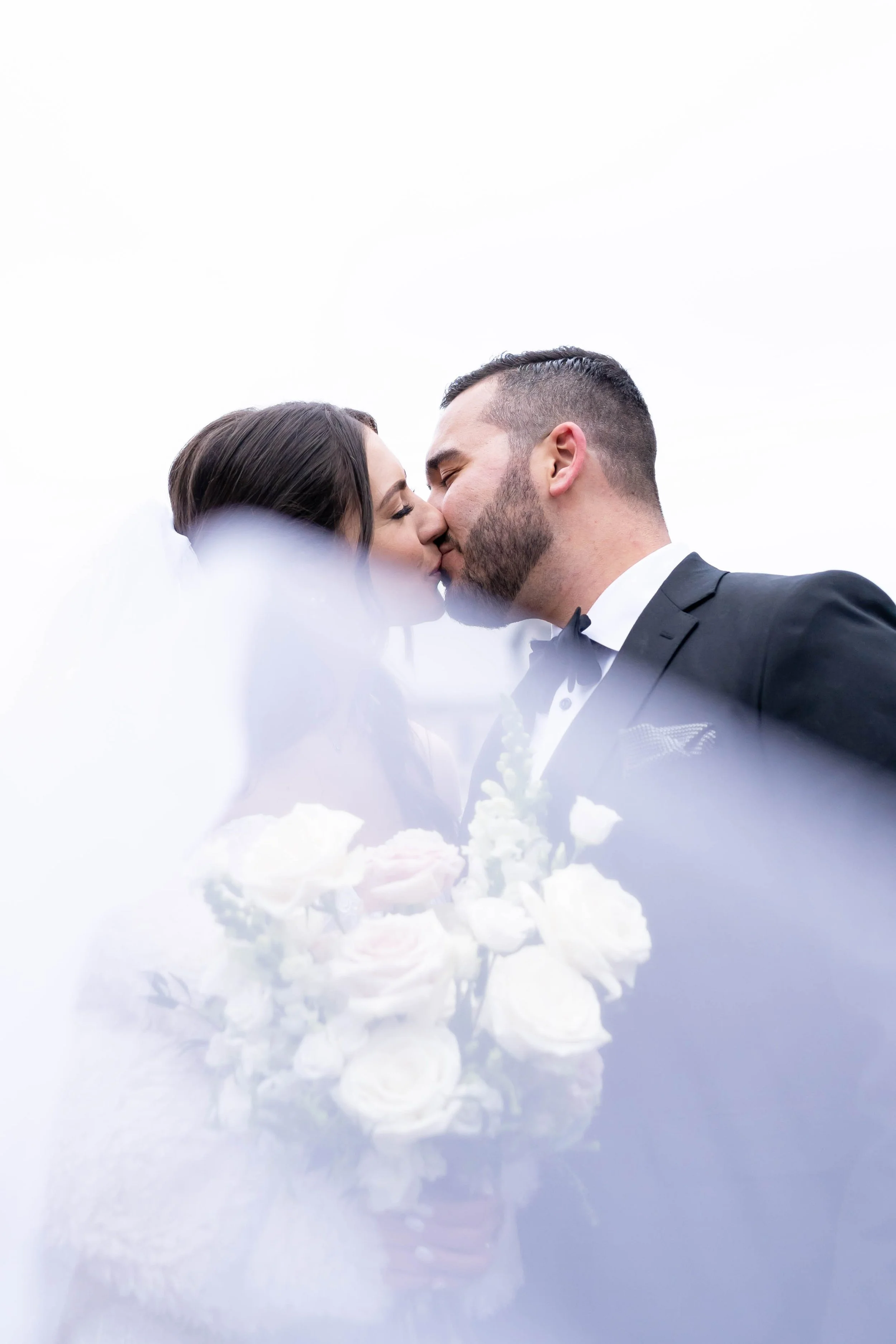 A couple in wedding attire sharing a kiss under an overcast sky, with rose petals and white flowers in the foreground.