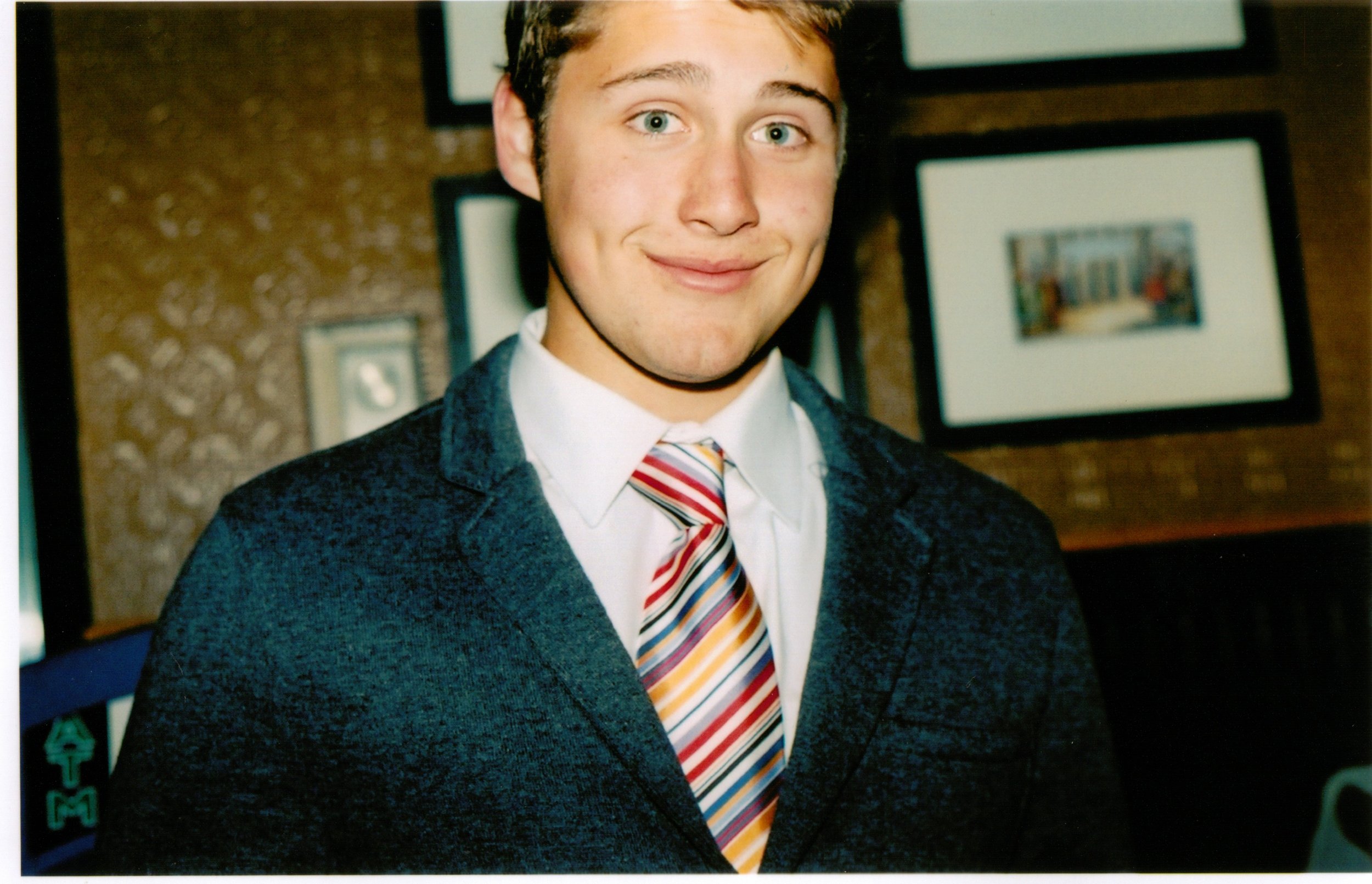 Young man with blue eyes, wearing a dark suit and a multicolored striped tie, smiling in front of framed pictures on a wall.