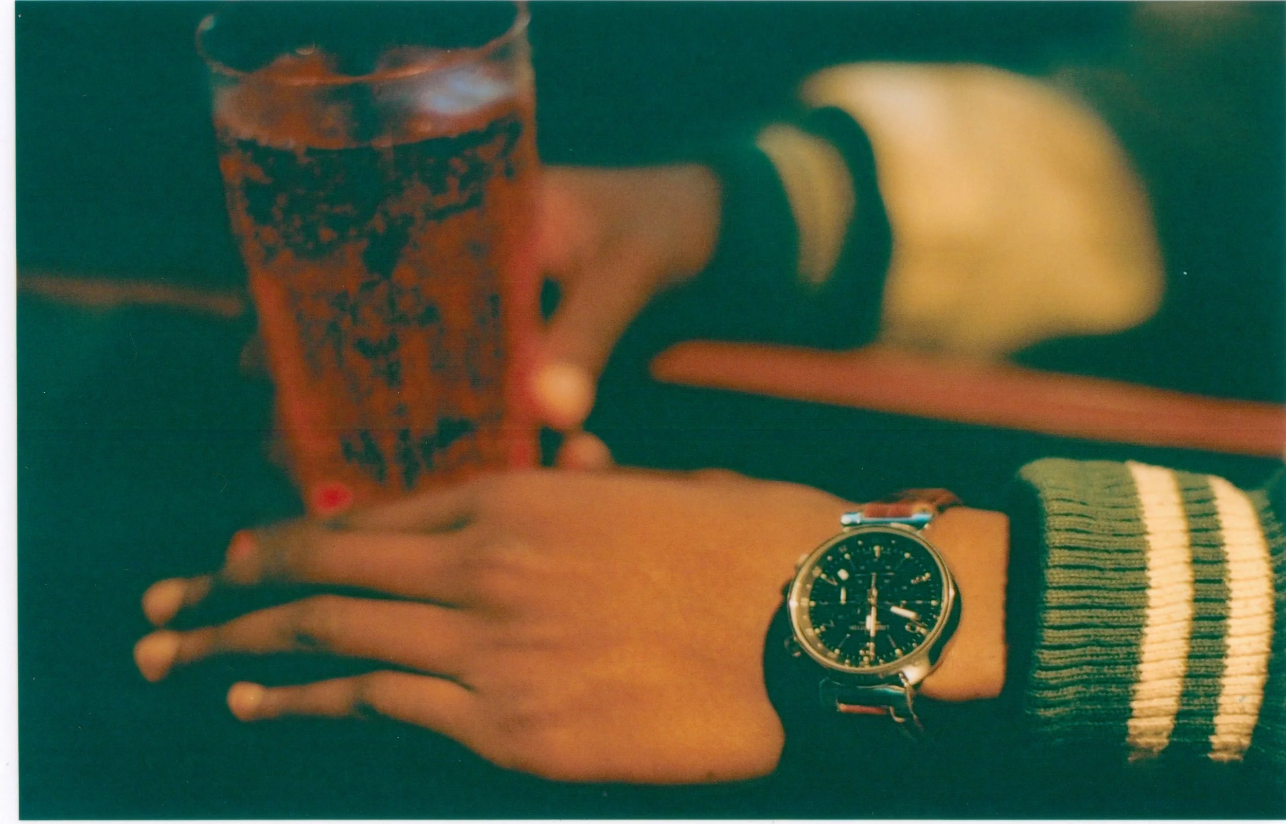 A person's hand wearing a striped sleeve and a watch resting on a wooden surface next to a striped sock, a glass of dark beverage, and a wristwatch. The photo appears to be a mirror selfie, with a wristwatch visible on the person's wrist.