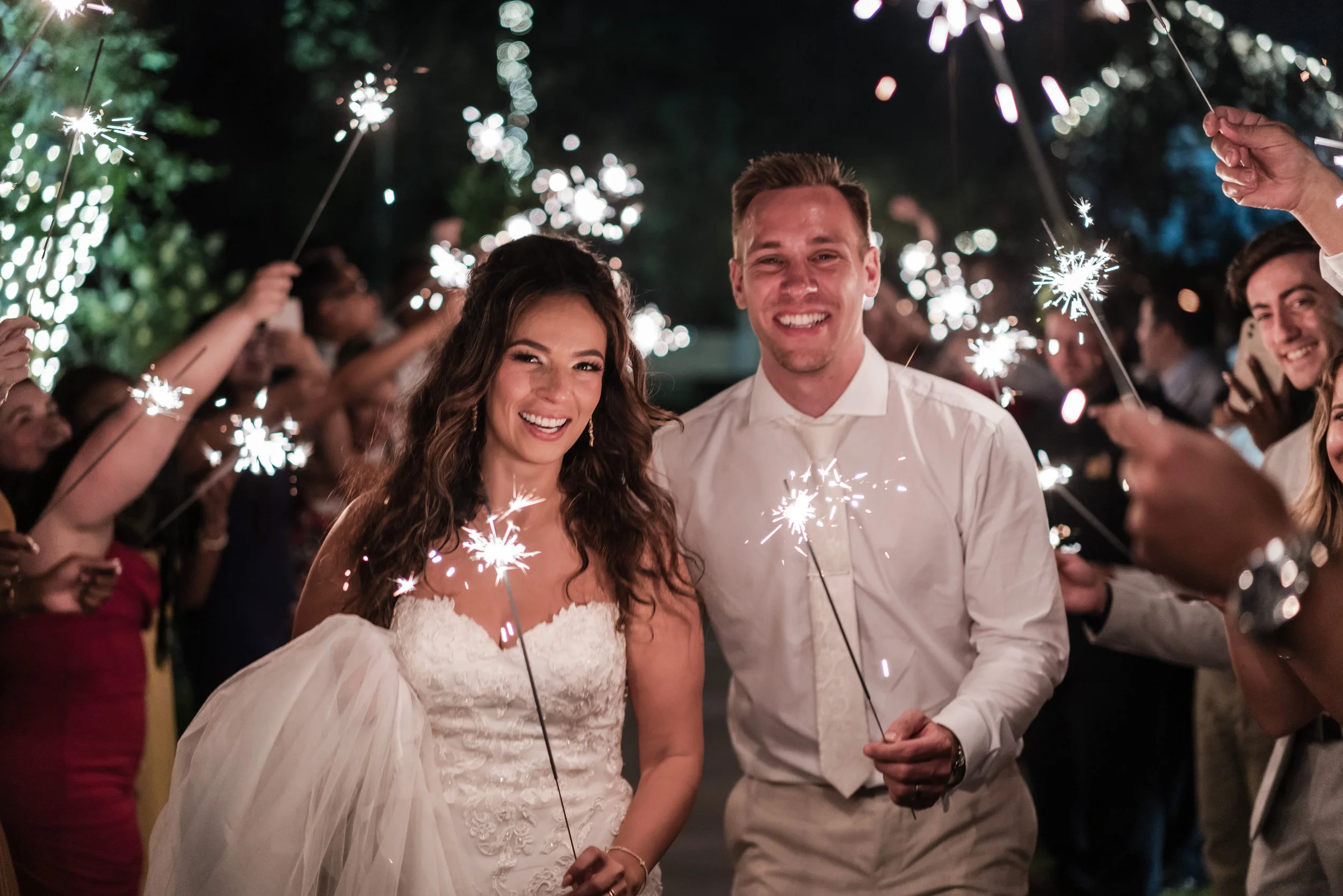 A bride and groom celebrating their wedding with sparklers at night, surrounded by guests also holding sparklers, smiling and enjoying the festive moment.