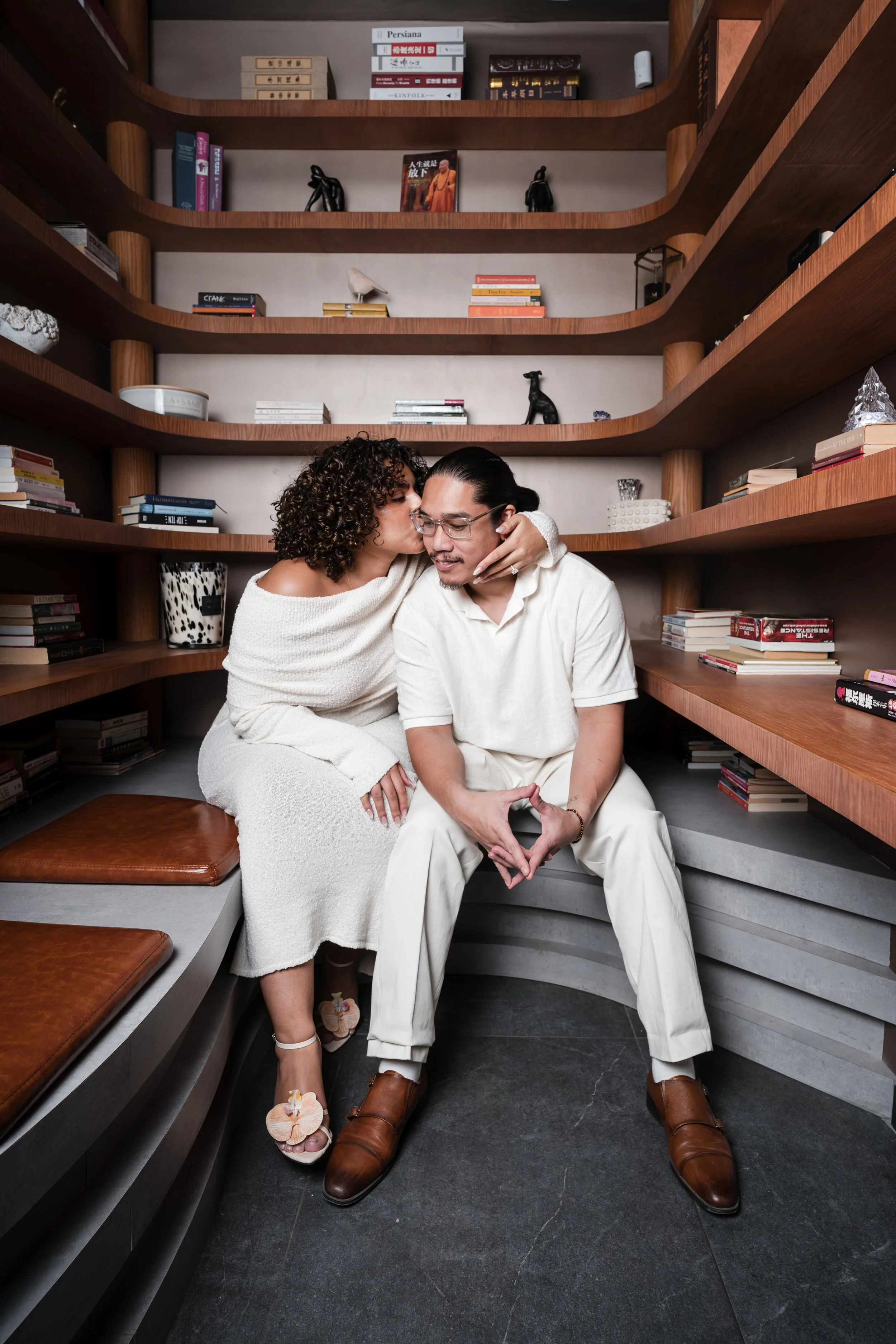 A woman and a man sitting close together on a bench surrounded by wooden bookshelves, with the woman whispering into the man's ear while he smiles. The woman has curly hair and is wearing a white dress, while the man has long hair tied back, glasses,