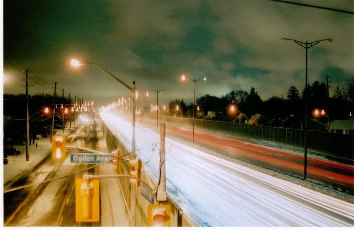 Nighttime street view with light trails from moving vehicles, streetlights, traffic signals, and Ogden Avenue sign.