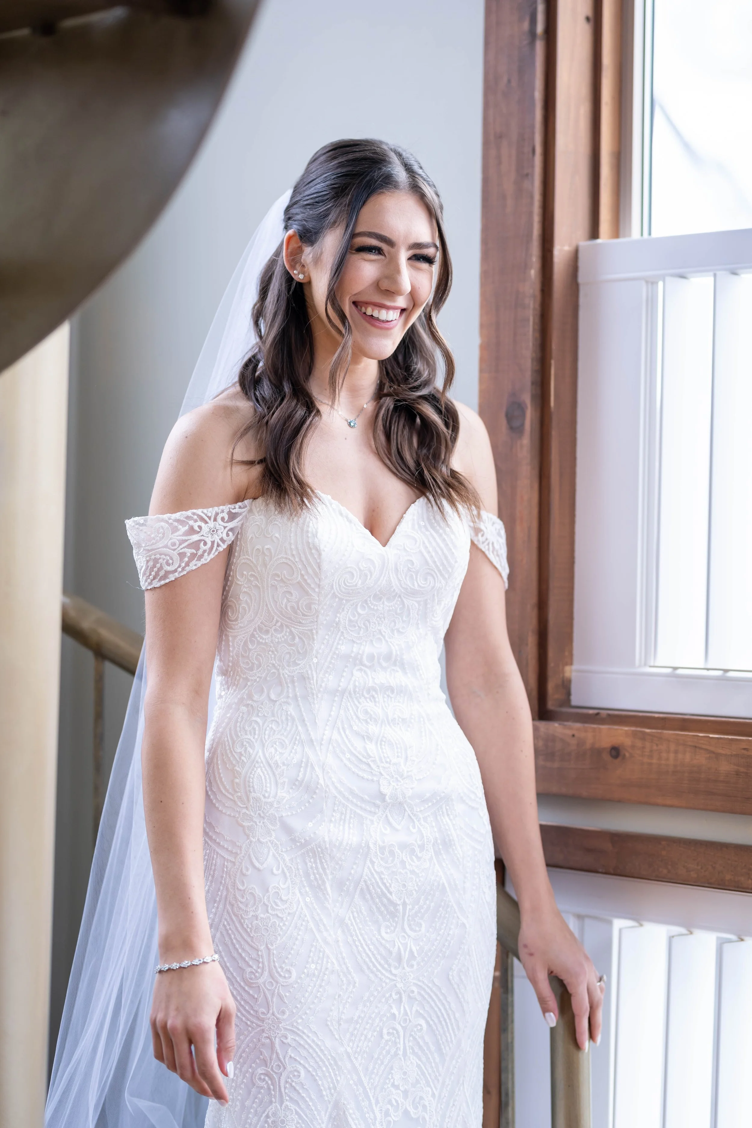 A young woman with dark brown hair in loose curls, wearing a white off-the-shoulder wedding dress with lace detailing, smiling and standing near a wooden window frame.