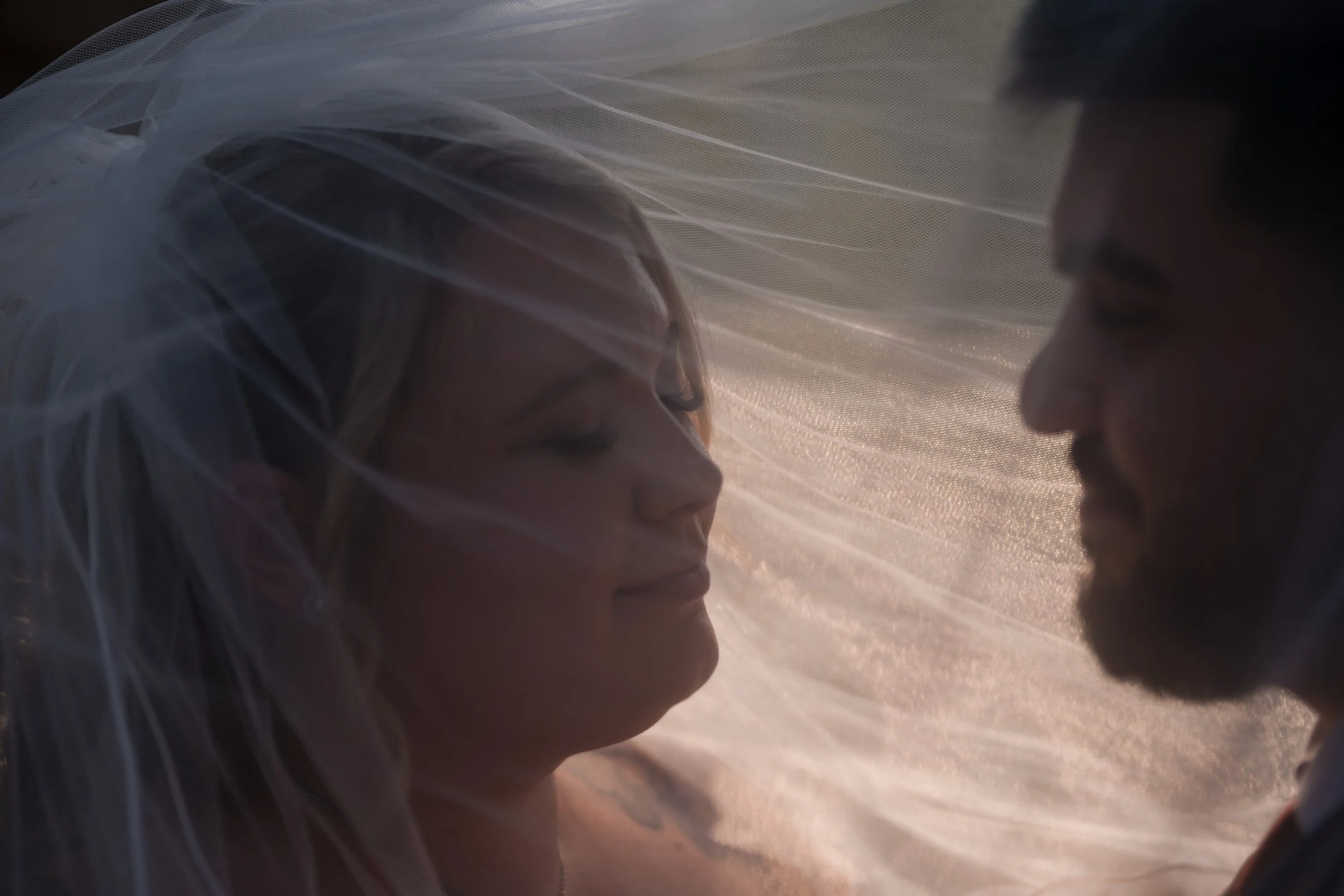 Close-up of a couple with their faces almost touching, backlit by warm light, with sheer fabric softly draped around them.