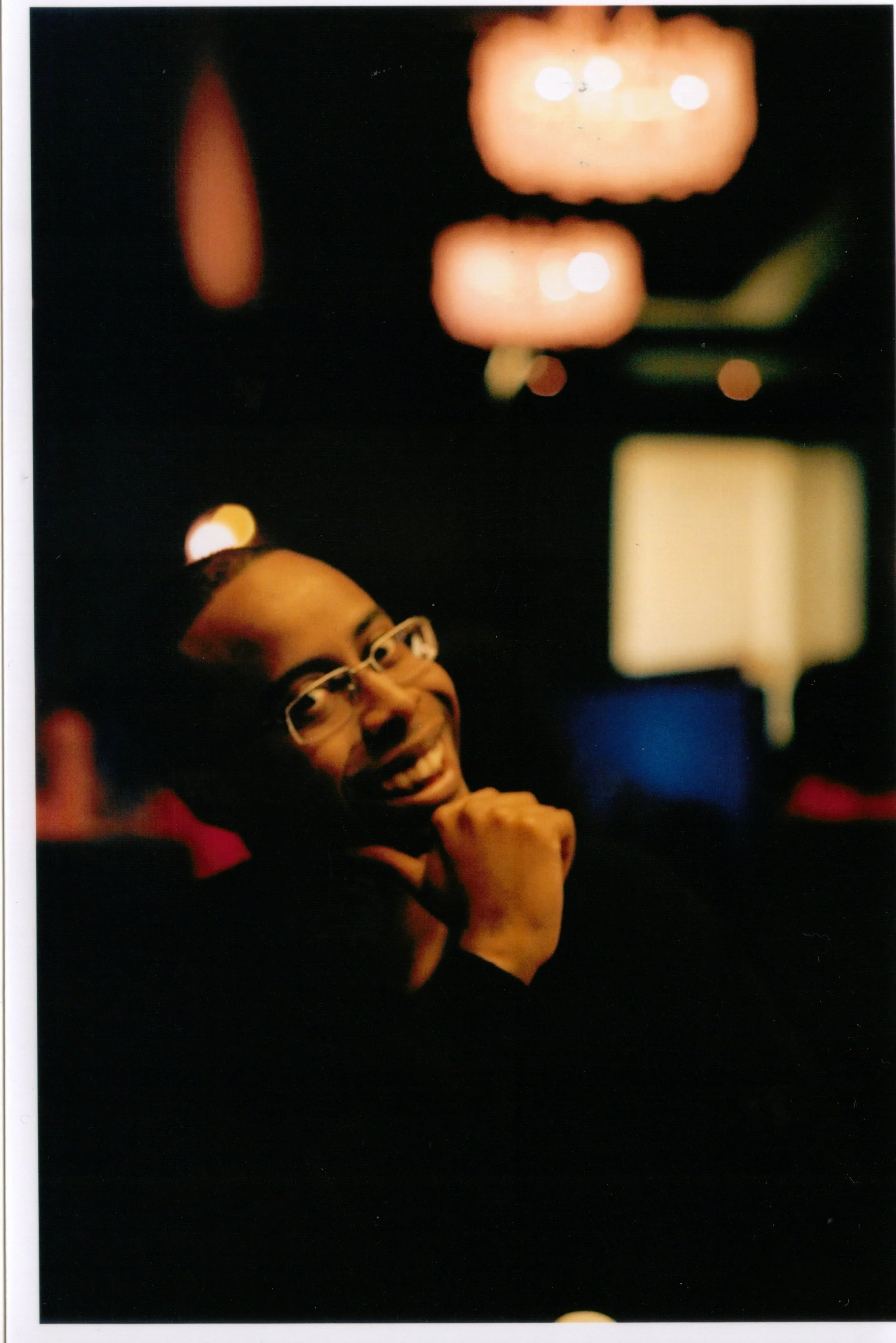 A woman with glasses and a bun hairstyle smiling and laughing in a dark setting with blurred, warm-colored lights in the background.