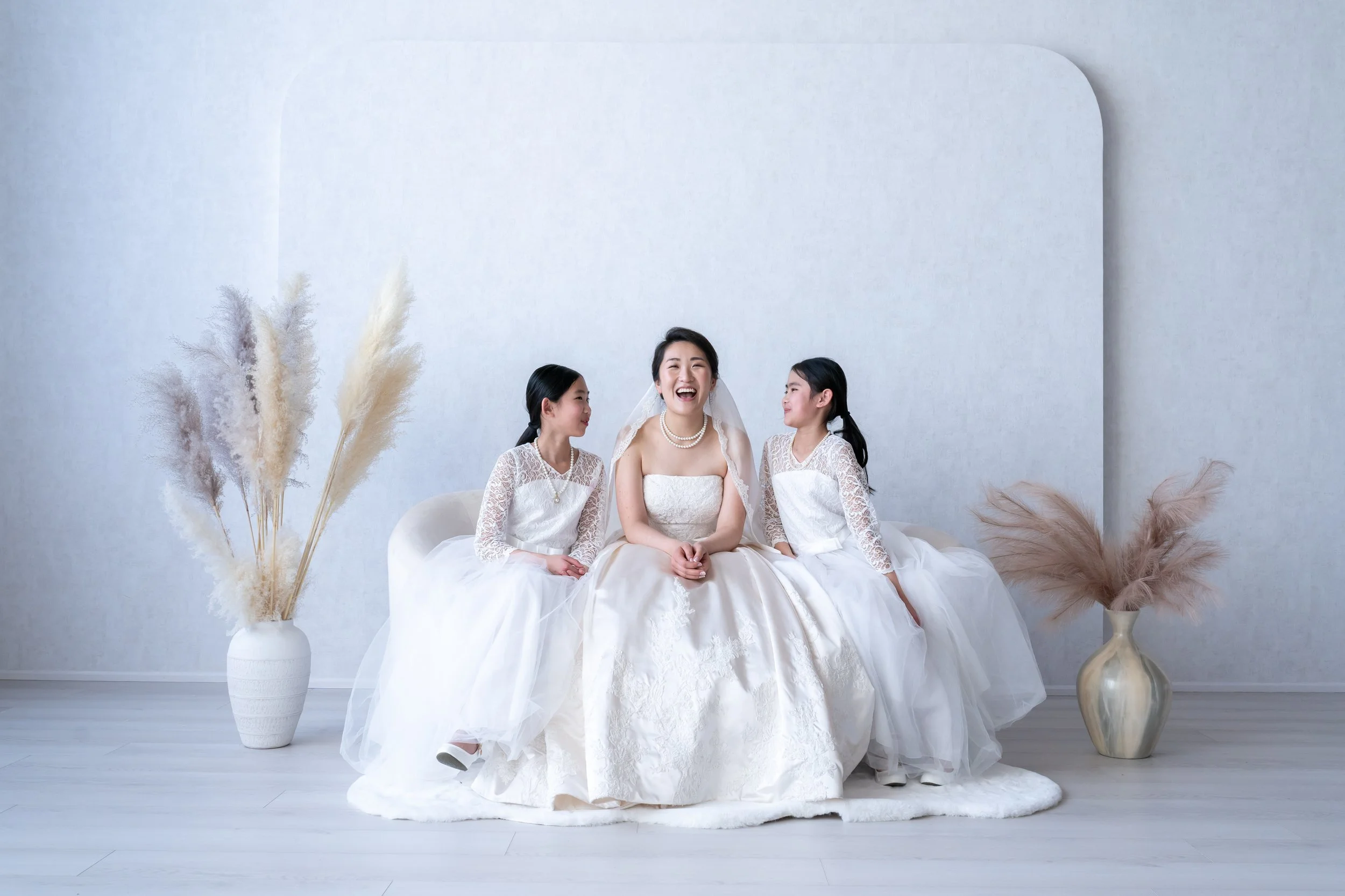 Three women in wedding dresses sitting on a white sofa in a minimalistic room with white walls and light-colored wooden floor, accompanied by large vases filled with pampas grass.