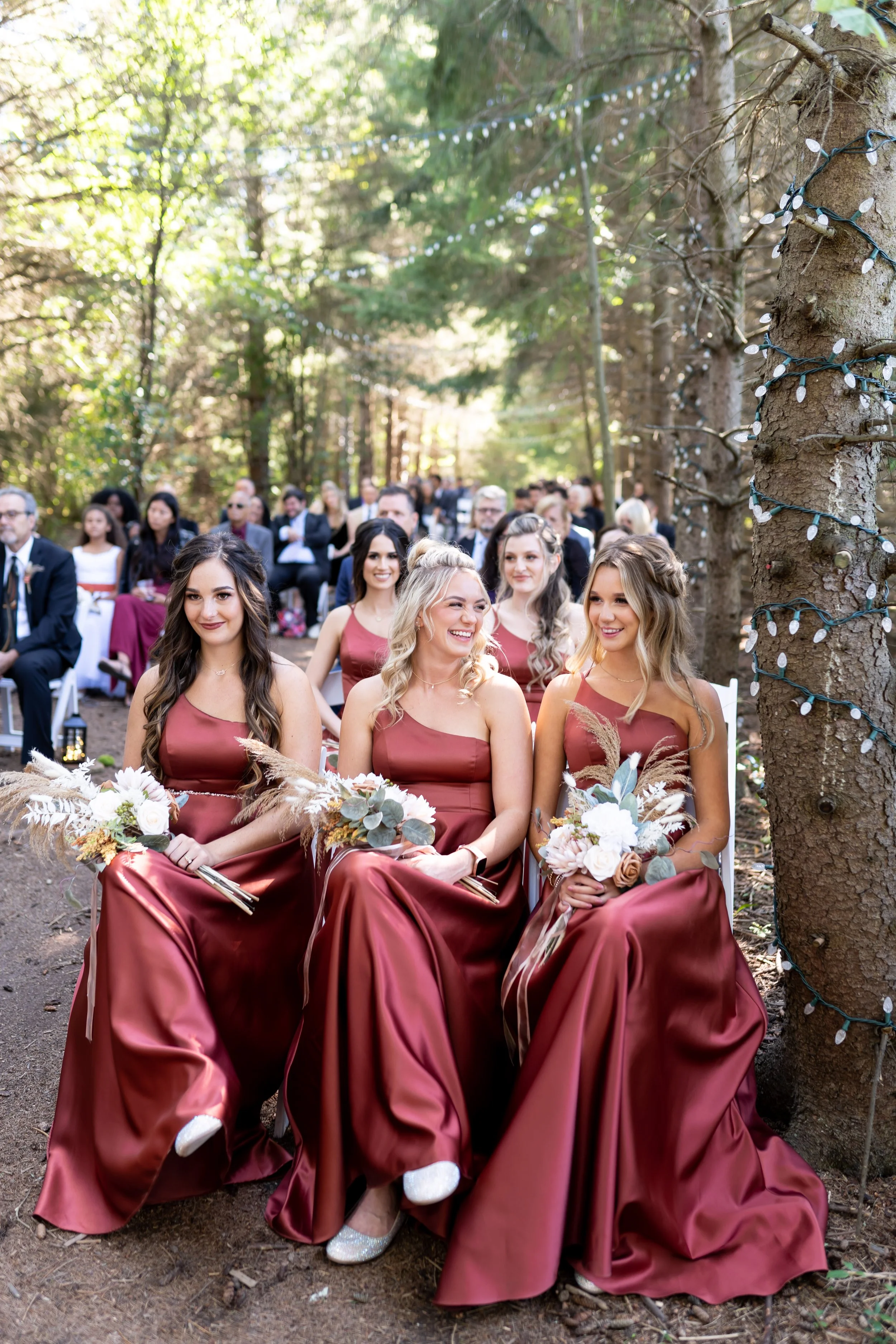 Bridal party sitting outdoors in a forest, wearing matching maroon satin dresses, holding bouquets, with string lights wrapped around nearby trees, at a wedding ceremony.