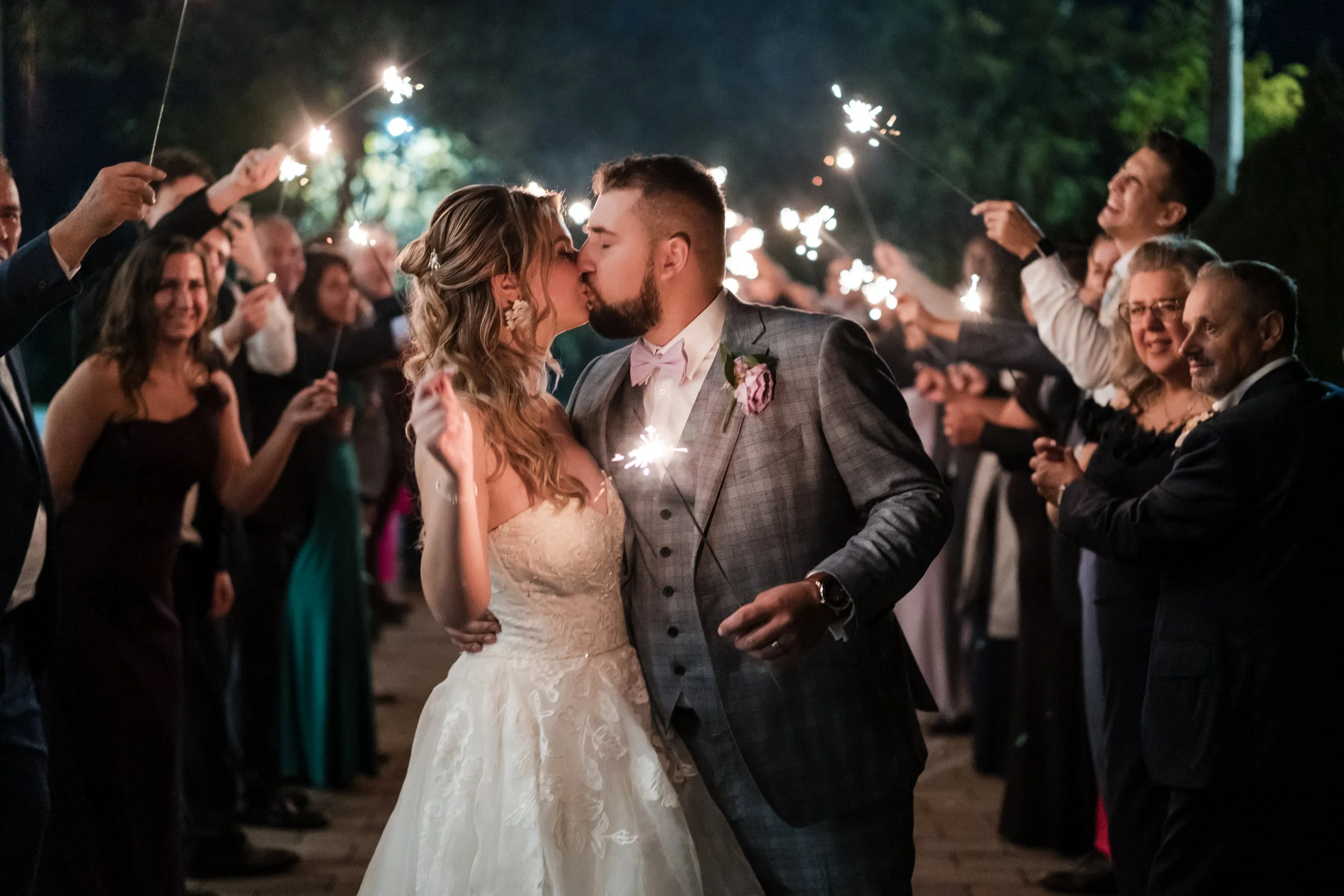 A bride and groom kiss surrounded by wedding guests holding sparklers at night.