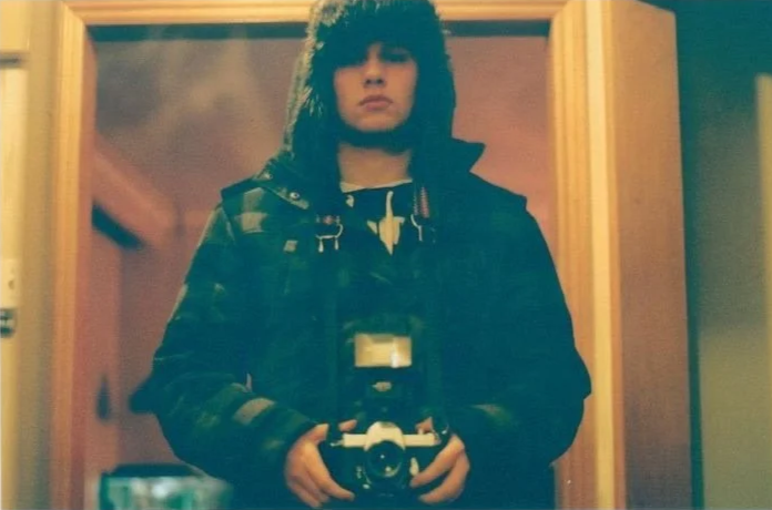 Joel taking a mirror selfie with a camera, wearing a dark jacket with a hood, standing in a room with a wooden-framed mirror and door.