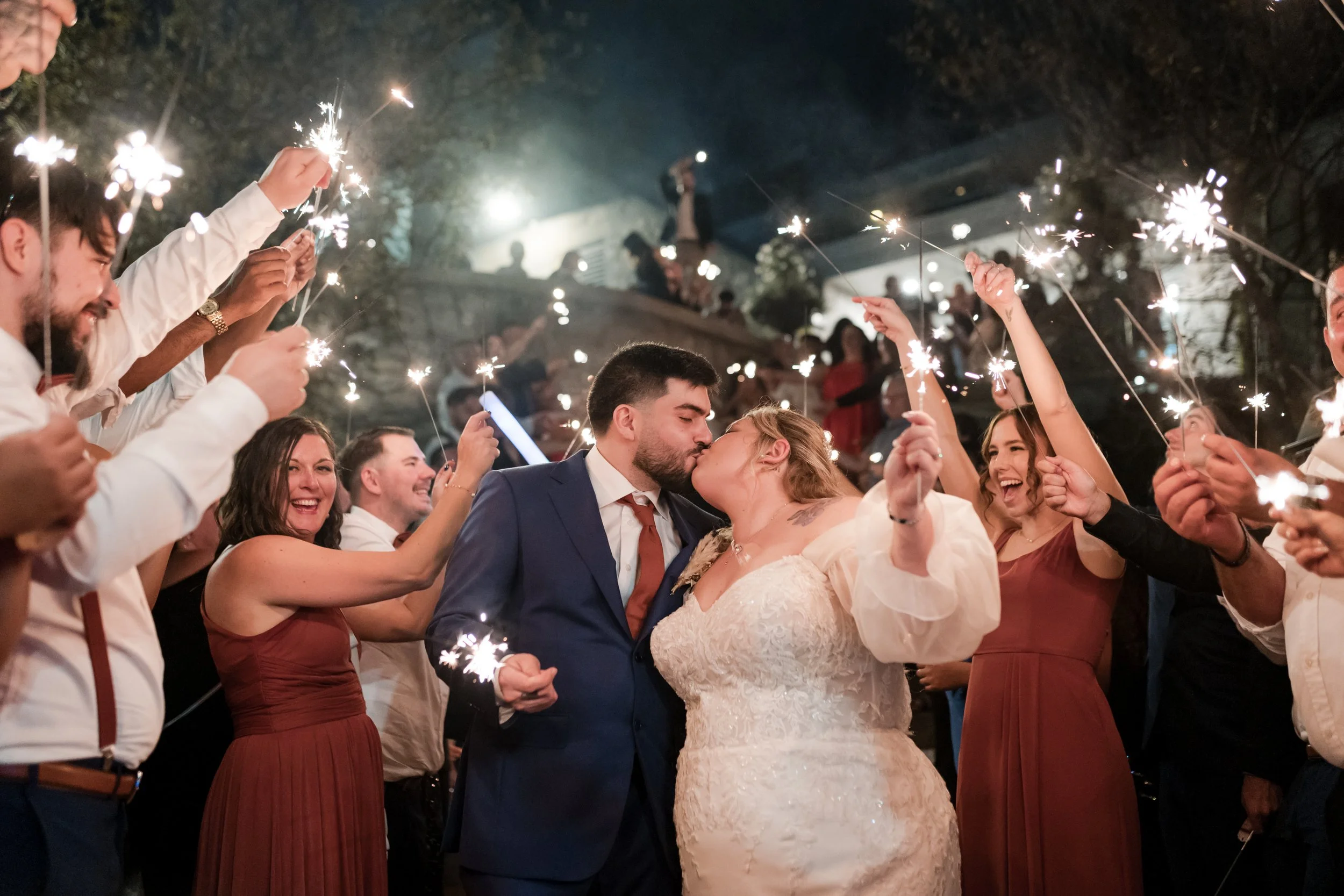 A bride and groom kiss in the center of a crowd at a nighttime outdoor celebration. The guests hold sparklers aloft, cheering and smiling around them.